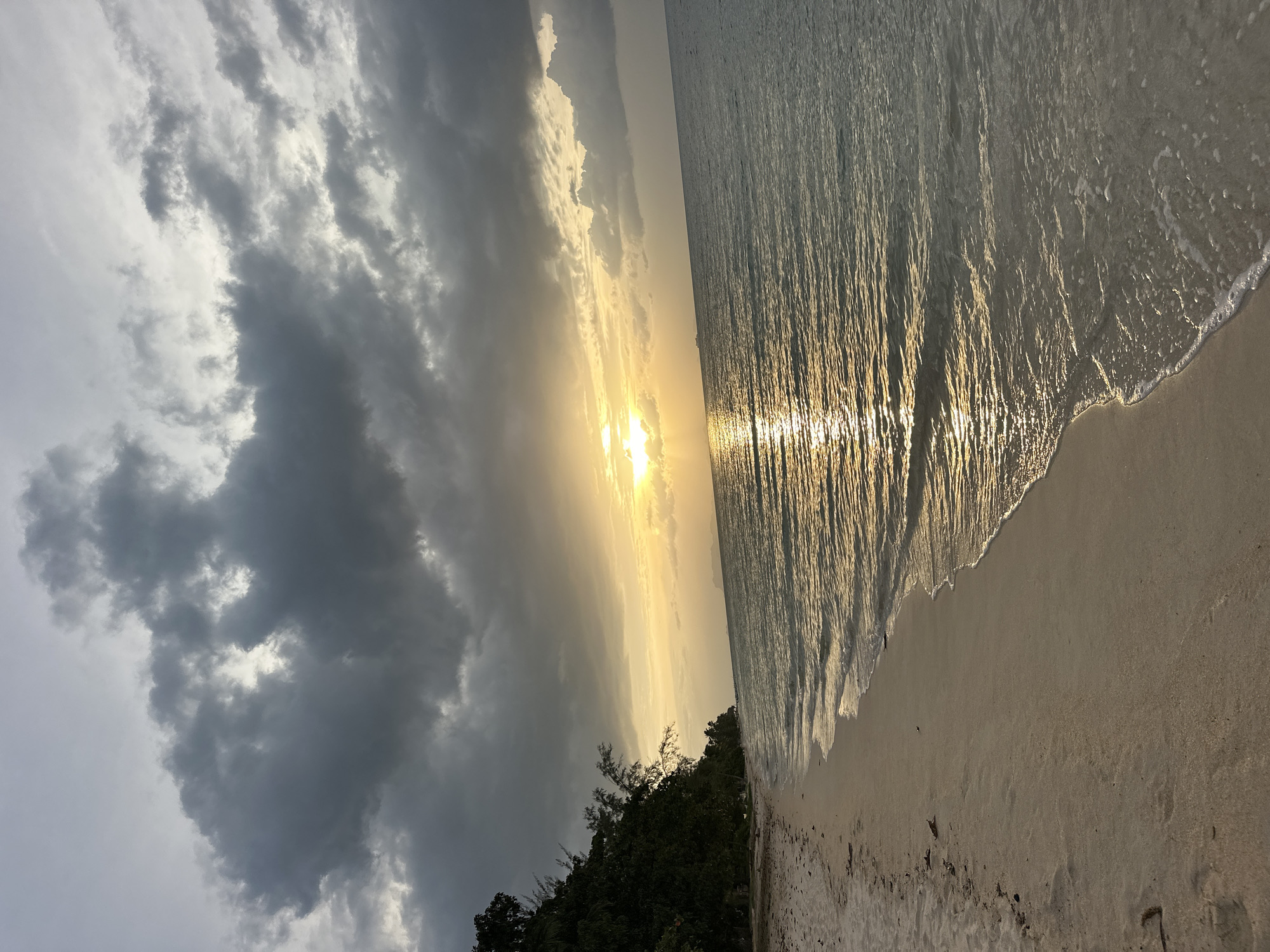 A serene beach scene unfolds in Ko Samui District, Thailand, as the sun begins its descent. The sky is a canvas of dramatic contrasts, with dark, heavy clouds dominating the upper portion, interspersed with rays of golden sunlight breaking through. These rays illuminate the water, creating a shimmering, reflective path from the horizon to the shore. The ocean itself is calm, with gentle waves lapping at the sandy beach.
To the left, a lush, green treeline marks the edge of the island, creating a natural boundary between the land and the sea. The sand is a light, neutral tone, with scattered debris from the tide. The overall impression is one of peaceful tranquility, with the dynamic sky adding a touch of drama to the otherwise still setting. There are no people or animals visible in the image, emphasizing the solitary beauty of the natural landscape.