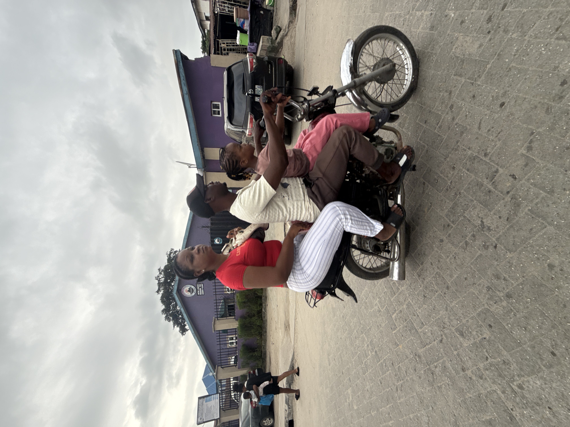 A family of three rides on a motorcycle on a paved road in Ado, Nigeria. The motorcycle is driven by a man wearing a dark baseball cap and a light-colored t-shirt. He is carrying a young girl in front of him, wearing a pink outfit. Behind the driver is a woman in a red t-shirt and striped pants. The background shows a purple building with a sign that reads "MFM LAGOS REGION TEN ADO-AJAH," indicating the location. To the left of the motorcycle, a woman in a black dress carries a baby, and a black car is parked on the side of the road. The sky is overcast with gray clouds.
