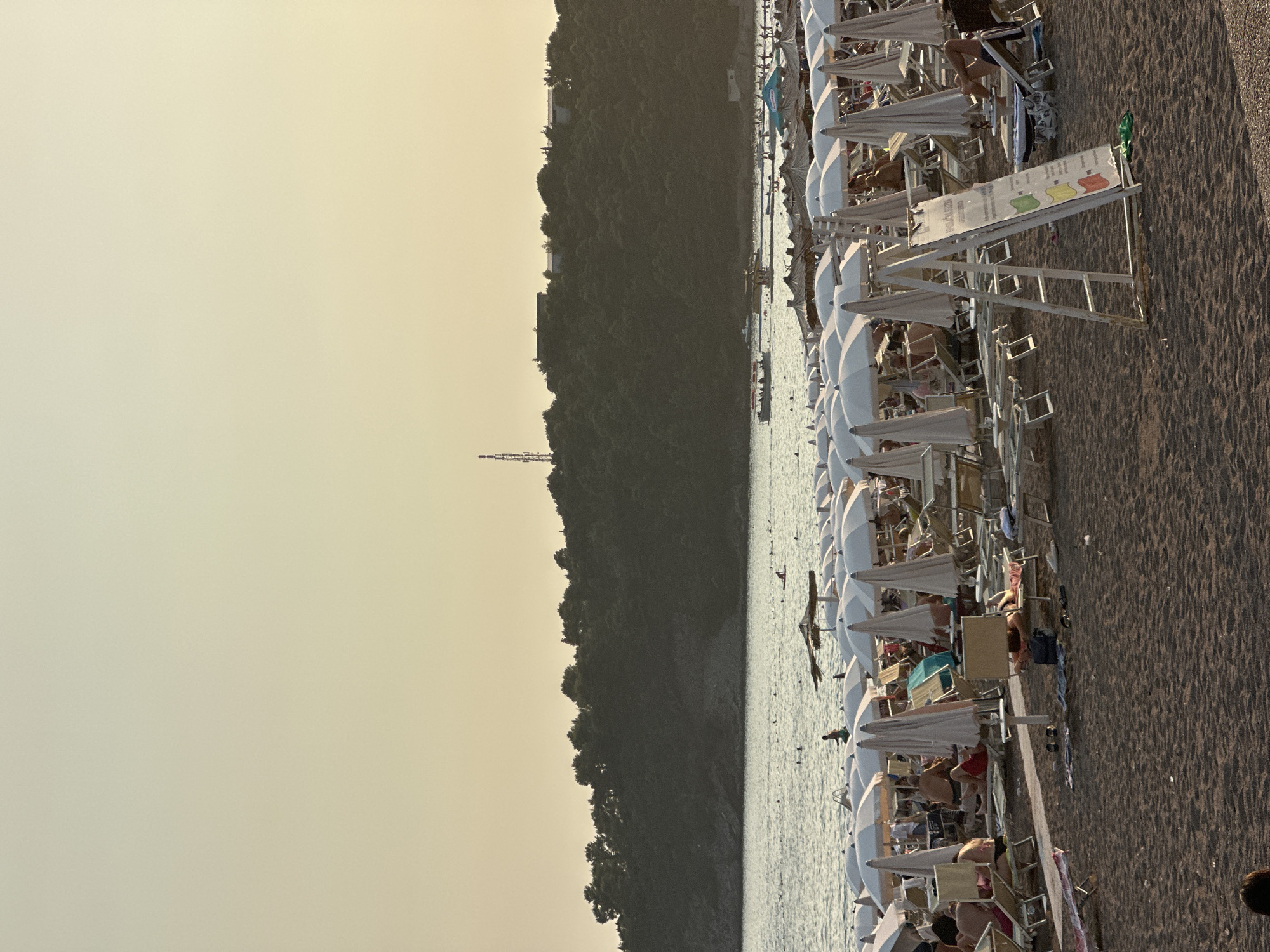 The image depicts a crowded beach in Boreti, Montenegro, during the daytime. The foreground is dominated by a sandy beach with numerous beach chairs and umbrellas, many of which are white and arranged in rows. Some people are visible lounging on chairs or walking on the beach. In the middle ground, the calm sea is dotted with swimmers and a few boats. The background features a densely forested hill with some buildings and a communication tower at the summit. The sky is a pale, hazy yellow, suggesting either early morning or late afternoon light. The overall scene conveys a sense of a busy tourist destination.
