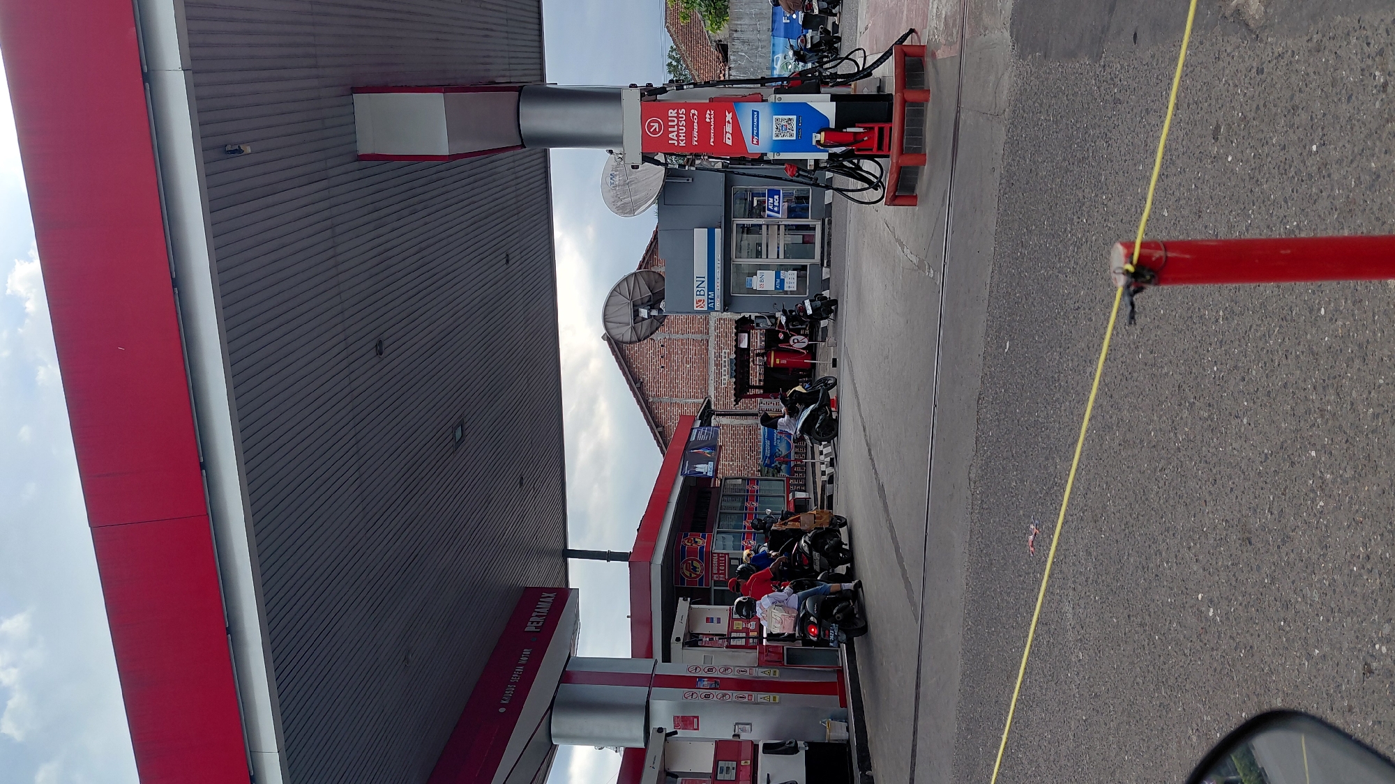 The image captures a scene at a Pertamina gas station, likely in Rejasari, Indonesia. Under a large red and white canopy, several motorcycles are parked and being refueled. Two individuals on a motorcycle are at the pump on the left, while another individual is refueling at a pump on the right. Behind the pumps, a brick building with satellite dishes is visible, and a BNI ATM booth is situated next to it. The area appears to be a typical urban or suburban setting, with asphalt ground and a cloudy sky. A yellow rope demarcates an area in the foreground.