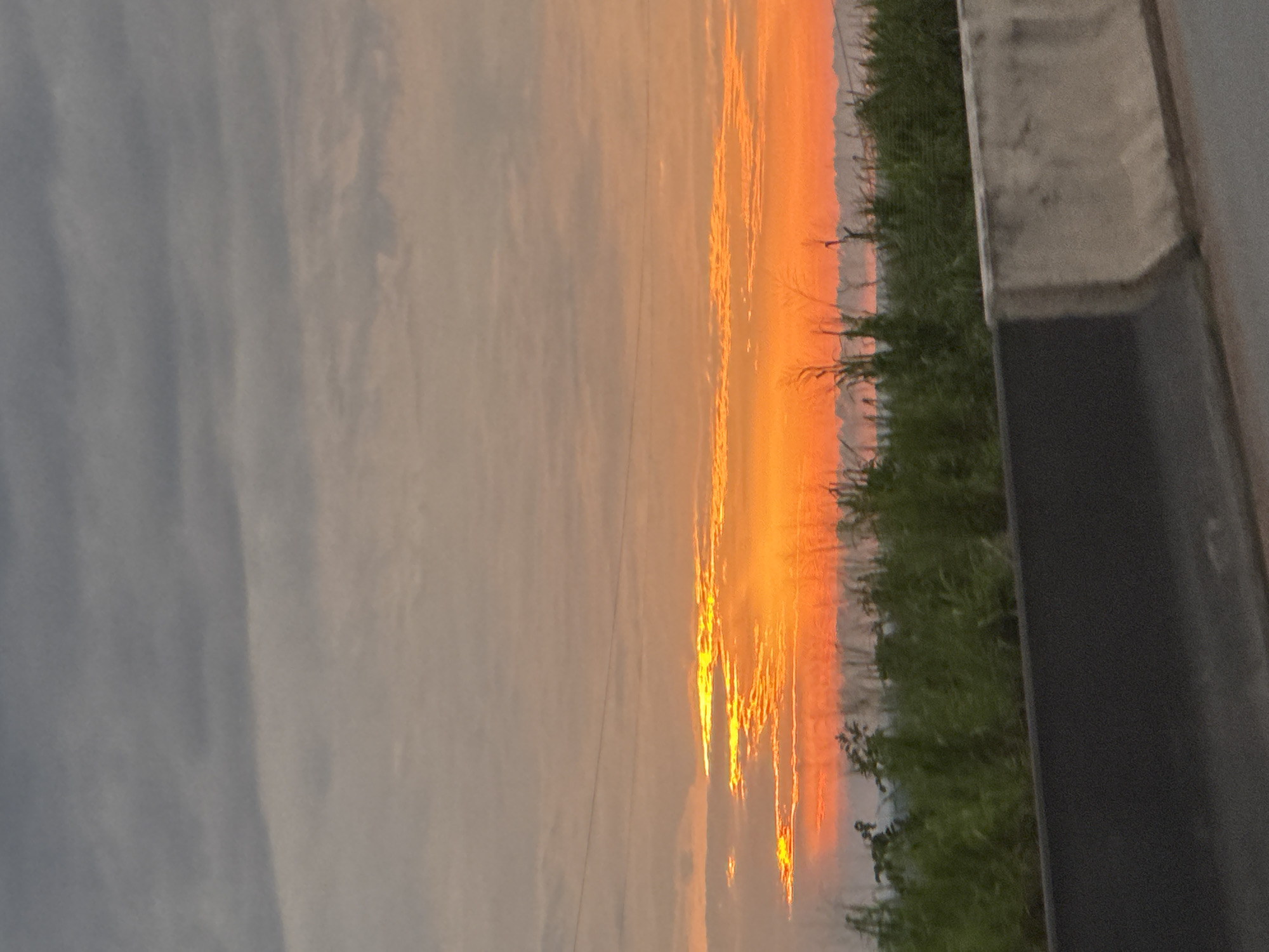 The image captures a vibrant sunset over Orifite, Nigeria. The sky is a canvas of warm oranges and yellows, with streaks of bright light piercing through the clouds. A thin layer of haze or fog hovers near the horizon, softening the edges of the distant landscape. In the foreground, lush green vegetation lines the roadside, with tall grasses and some scattered foliage. A concrete barrier, typical of highway infrastructure, is visible in the lower portion of the frame, separating the viewer from the natural scene beyond. The overall impression is one of natural beauty as day transitions into night.