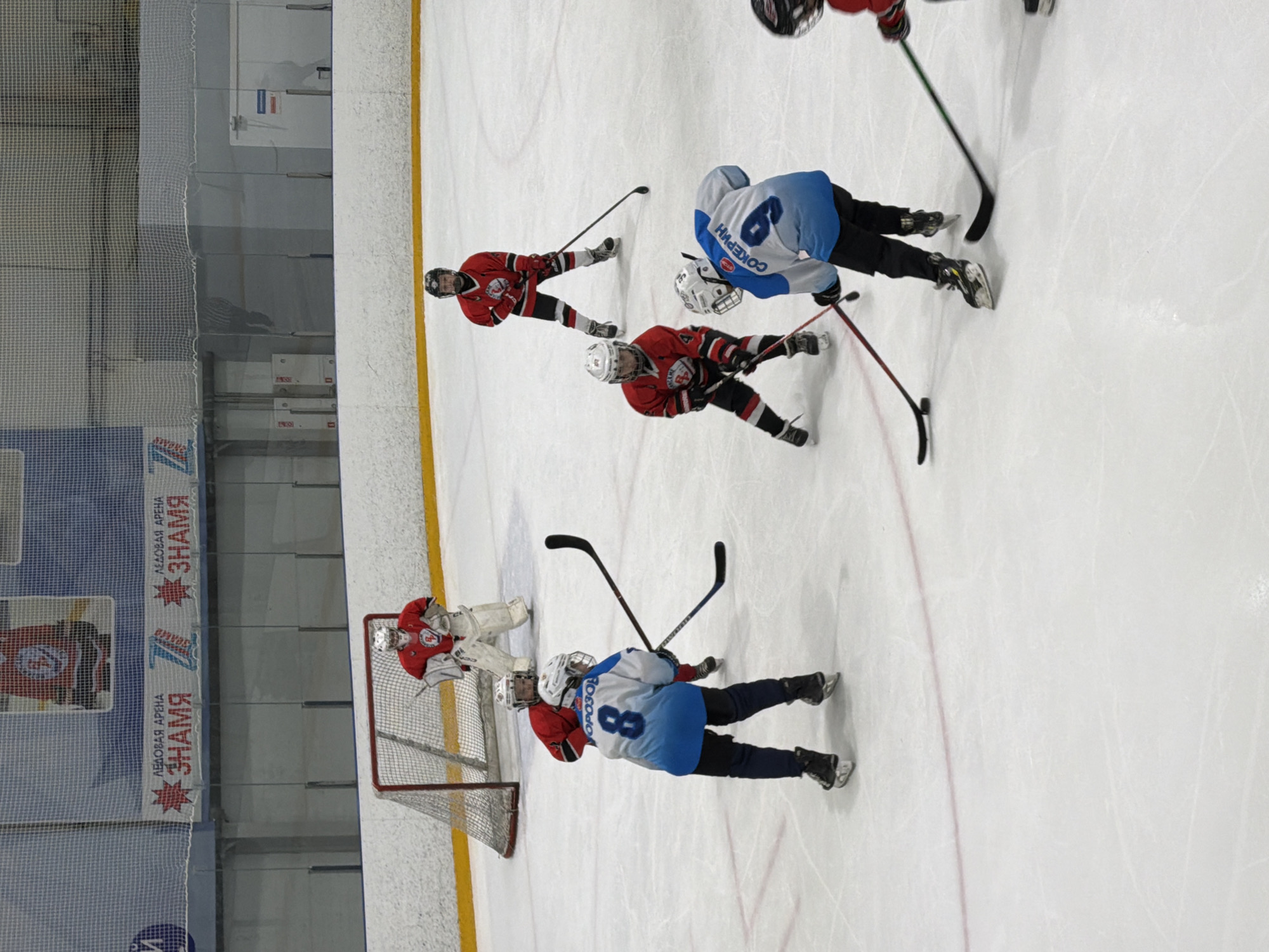 An aerial view captures an active ice hockey game or practice session inside an arena. The image is rotated 90 degrees clockwise, showing players on a bright white ice surface with visible yellow and red lines.
The scene features several ice hockey players from two teams. One team wears red jerseys with black accents, while the opposing team wears light blue and white jerseys. Near the center of the frame, two players in red jerseys are positioned, appearing to face towards the goal. Close by, a player in a light blue jersey, identified by the name "СОКЕРИН" (Sokerin) above the number 6 on their back, is engaged in play.
Towards the right side of the rotated image (the original bottom), a hockey net stands, guarded by a goalie in a red and white uniform. Another player in a light blue jersey, with the name "МОРОЗОВ" (Morozov) above the number 8, is positioned near the net. All players are equipped with hockey sticks, helmets, and skates.
The background reveals the arena's structure, including protective netting and walls. Banners are visible along the arena wall, providing clear location cues. Text on the banners reads "ЛЕДОВАЯ АРЕНА" (Ice Arena) and "ЗНАМЯ" (Znamya). Although partially obscured, another banner likely indicates "ВОТКИНСК" (Votkinsk), confirming the setting as the Ice Arena Znamya in Votkinsk, Russia. The focus is on the action of the game, with players demonstrating various postures of active play.