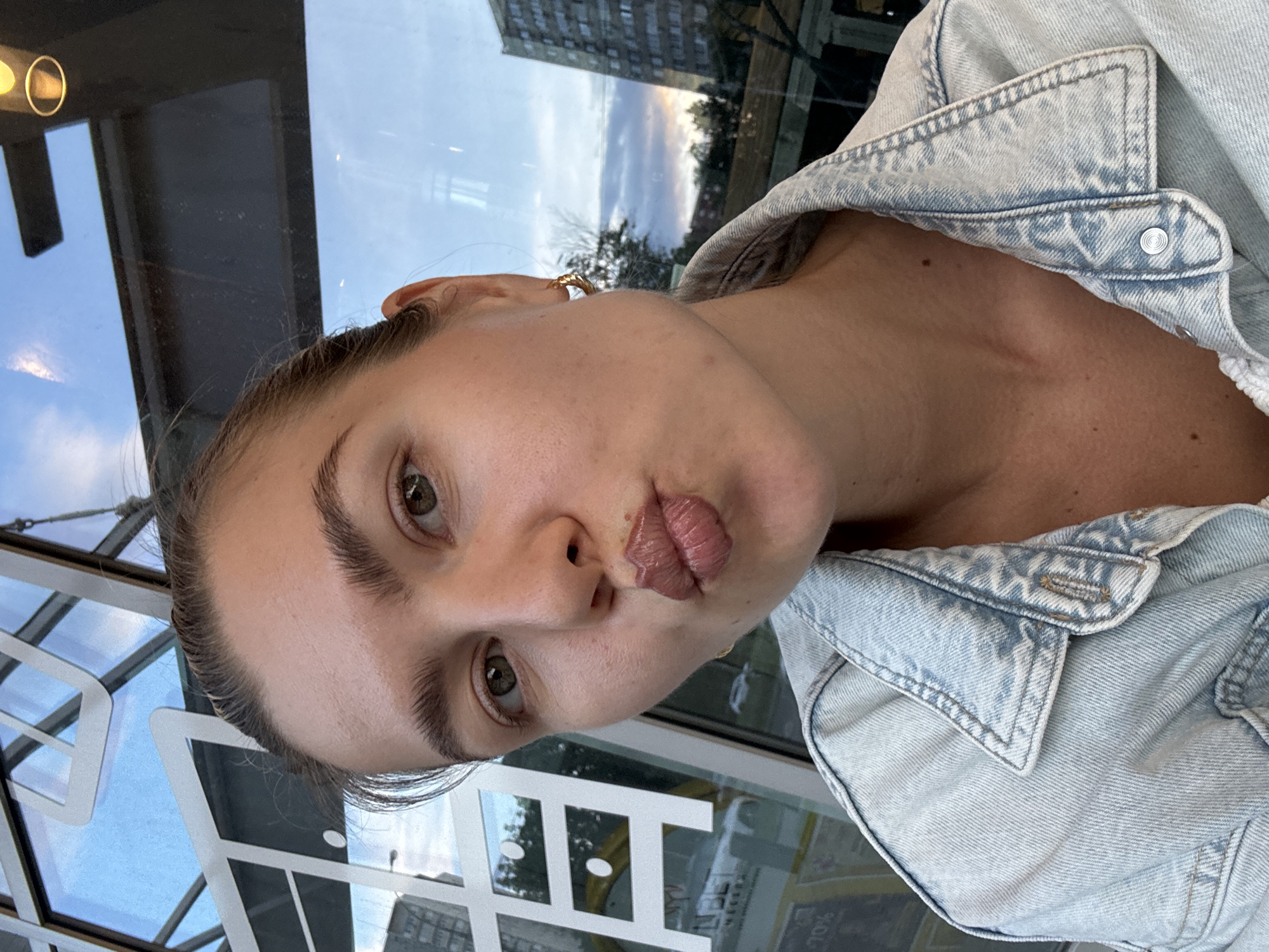 A young woman, with her hair pulled back, is taking a selfie in front of a window. She is wearing a light blue denim jacket and a gold hoop earring in her left ear. Her lips are pursed in a kiss pose, and she is looking directly at the camera. The window behind her reflects the sky and buildings, suggesting an outdoor or indoor urban setting. A large white graphic element, possibly part of a sign or building facade, is visible on the left side of the frame.
