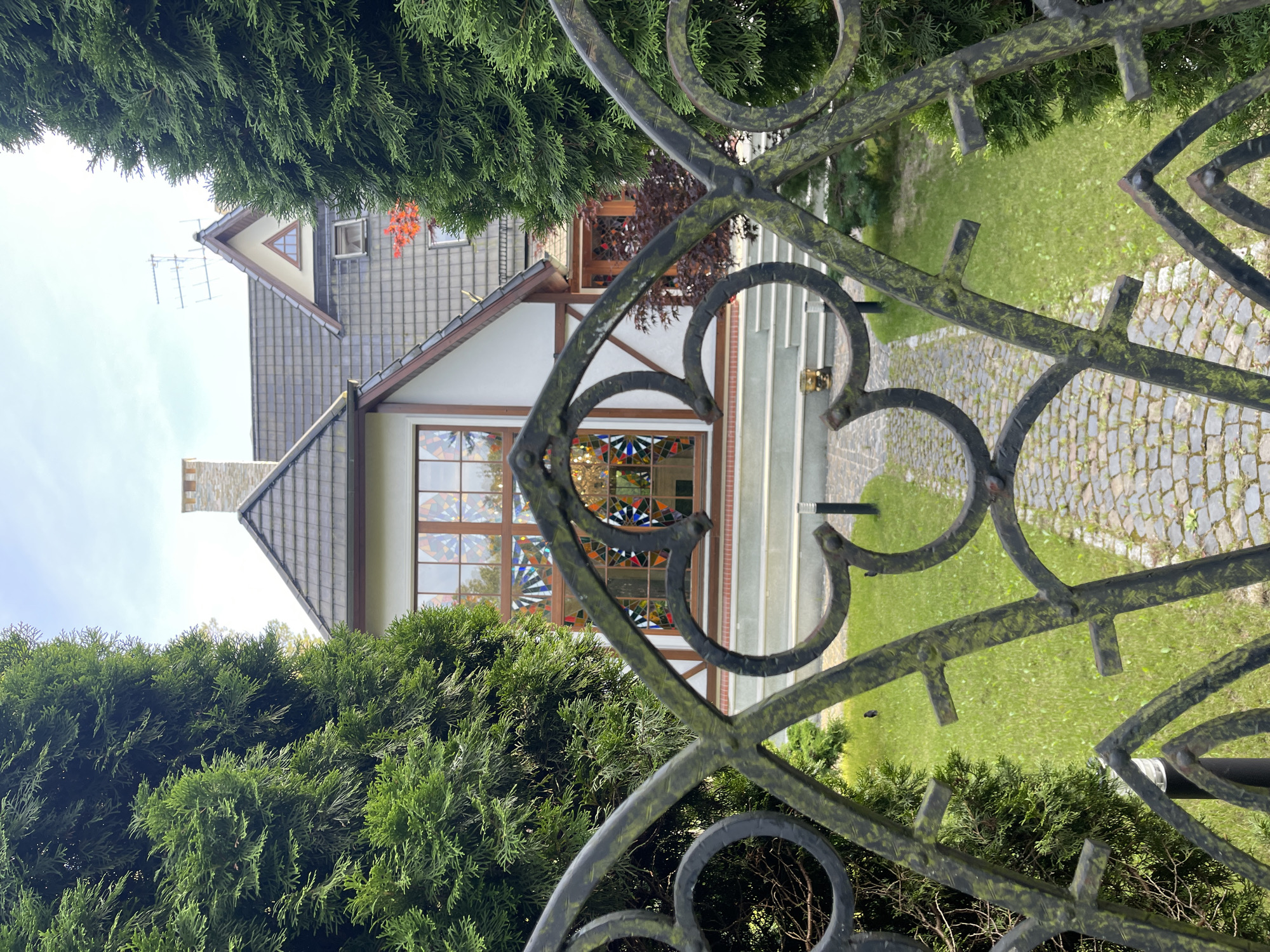 The image captures a view through an ornate, dark metal gate, which is partially covered in green moss. Beyond the gate, a house with a steeply pitched, dark gray tiled roof is visible. The house features white walls with some dark trim and timber framing accents. A prominent stained-glass window with a geometric pattern in shades of blue, red, and yellow is the centerpiece of the facade. The window is framed by wood. A cobblestone pathway leads up to a set of white stairs that ascend to the entrance of the house. Flanking the pathway and entrance are well-maintained green lawns and lush, dark green evergreen trees. The scene appears to be set in Svetlogorsk, Russia, on a bright day with a clear sky. The overall impression is one of a well-kept, possibly residential property with distinctive architectural features.