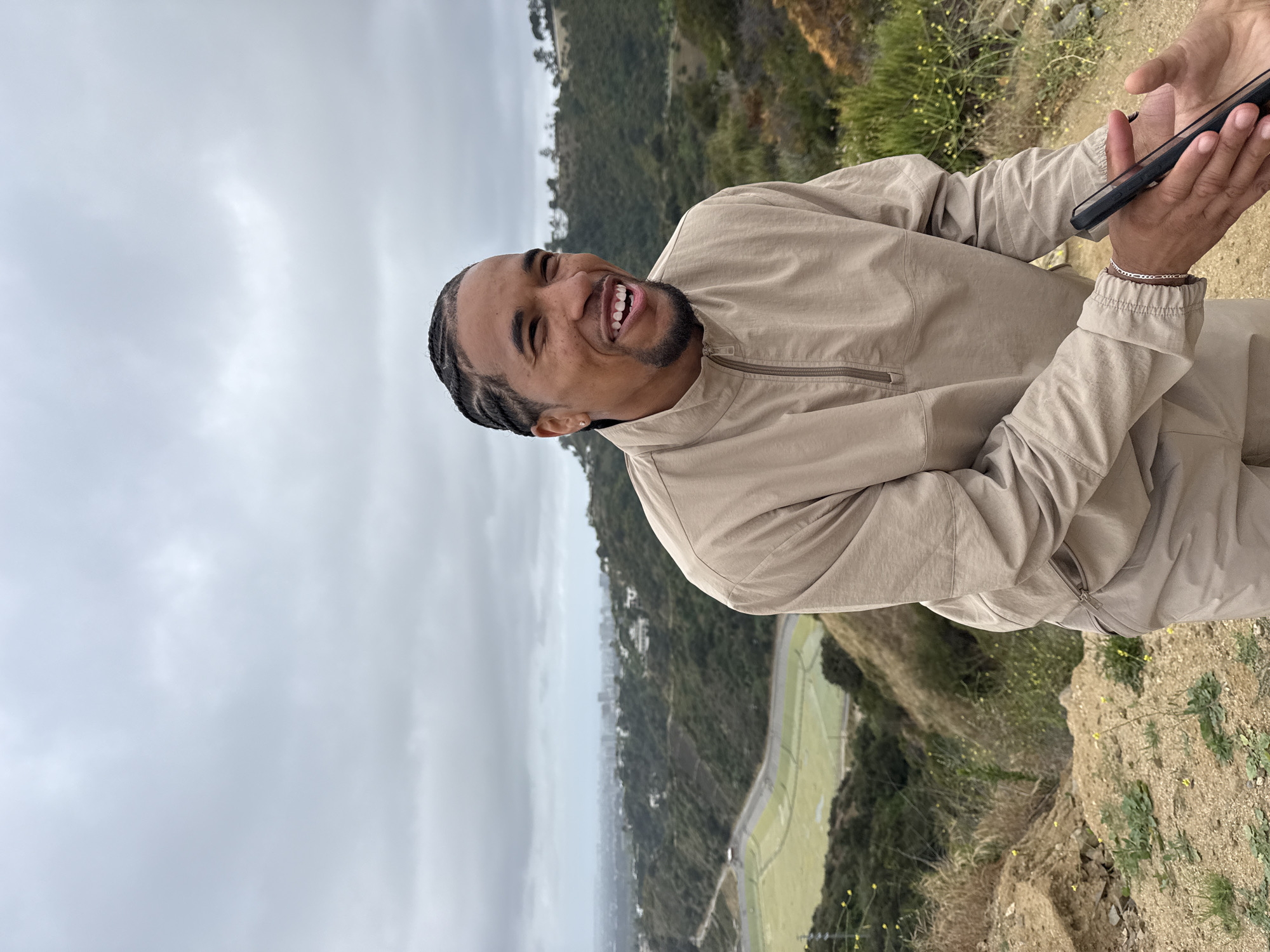 A man stands smiling on a hillside above the city of Los Angeles, CA. The location is likely a part of Griffith Park near the Hollywood Bowl, given the terrain and visible landmarks. He is holding a smartphone up as if taking a selfie or recording a video. He is dressed in tan clothing. The background includes trees, other parts of the hillside, a park with light green grass, and the city in the distance. The sky is overcast.
