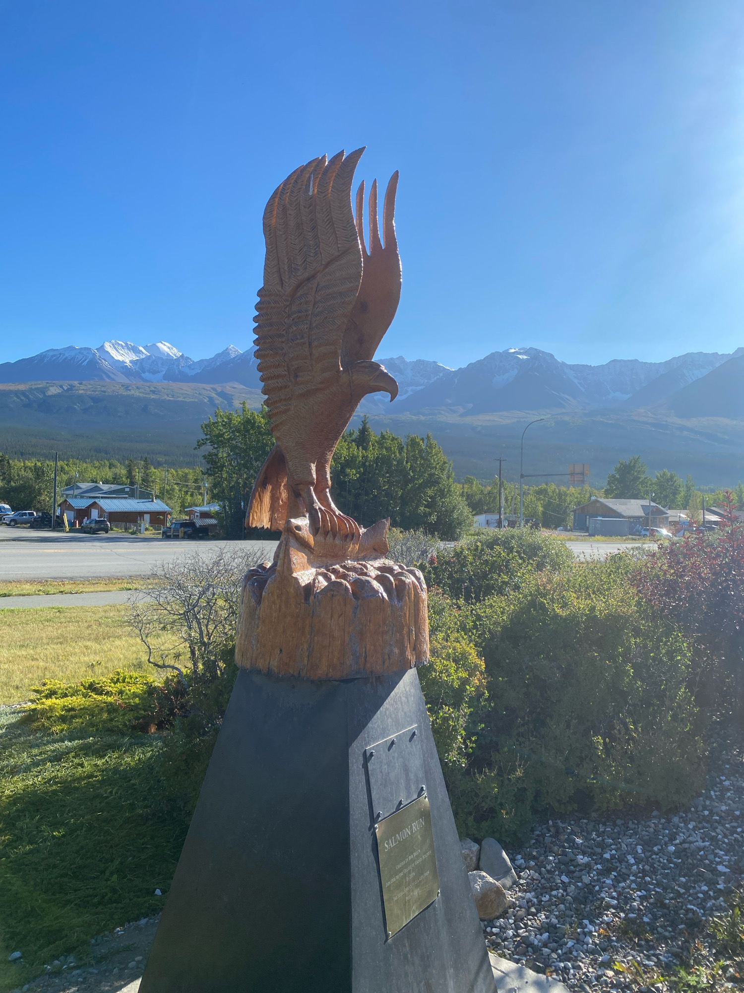 A carved wooden eagle sculpture is positioned in the foreground, its wings raised as if in flight, on a dark, conical pedestal. The pedestal has a plaque that reads "SALMON RUN." Behind the sculpture, the town of Haines Junction, Canada, is visible, with buildings, trees, and a road. In the background, a majestic mountain range with snow-capped peaks stretches across the horizon under a clear blue sky. The sun is shining brightly, casting long shadows. The environment appears to be a sunny day in a mountainous region.