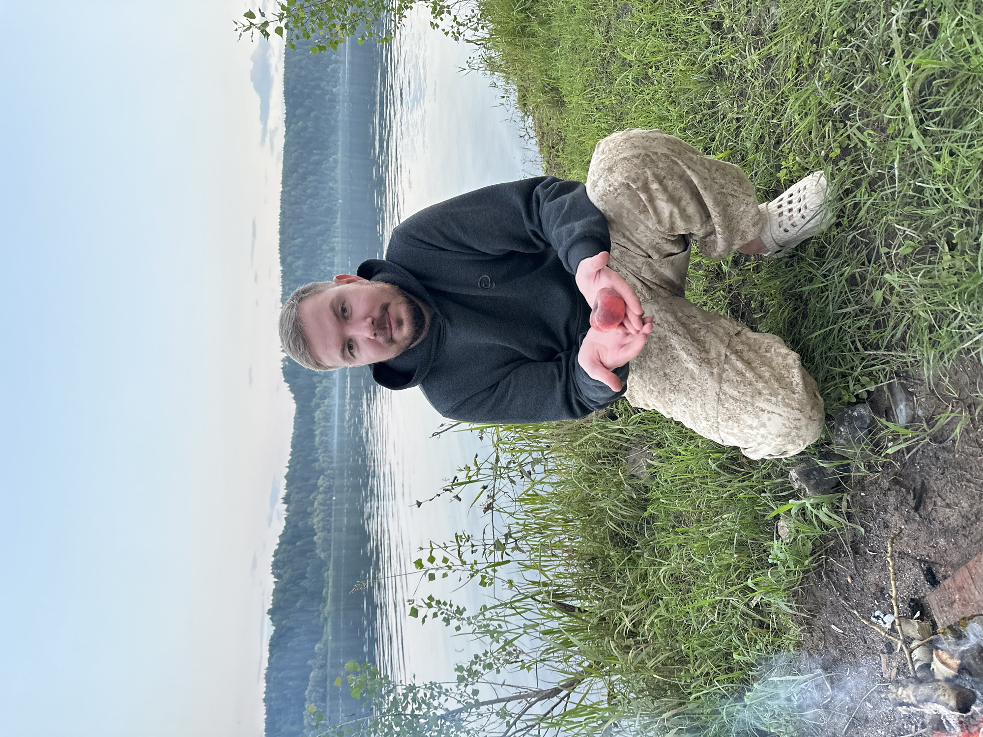 A man with short brown hair and a beard kneels on a grassy bank beside a wide, calm river. He is wearing a dark gray hooded sweatshirt and camouflage-patterned pants. In his open palms, he holds a small, red, heart-shaped object, possibly a fruit or a decorative item. In the background, a dense, dark green forest lines the opposite bank of the river, and the sky is a pale blue with a few wispy clouds. To the left of the man, a small campfire with visible smoke is present on the ground. The location is described as Noviy, Russia.