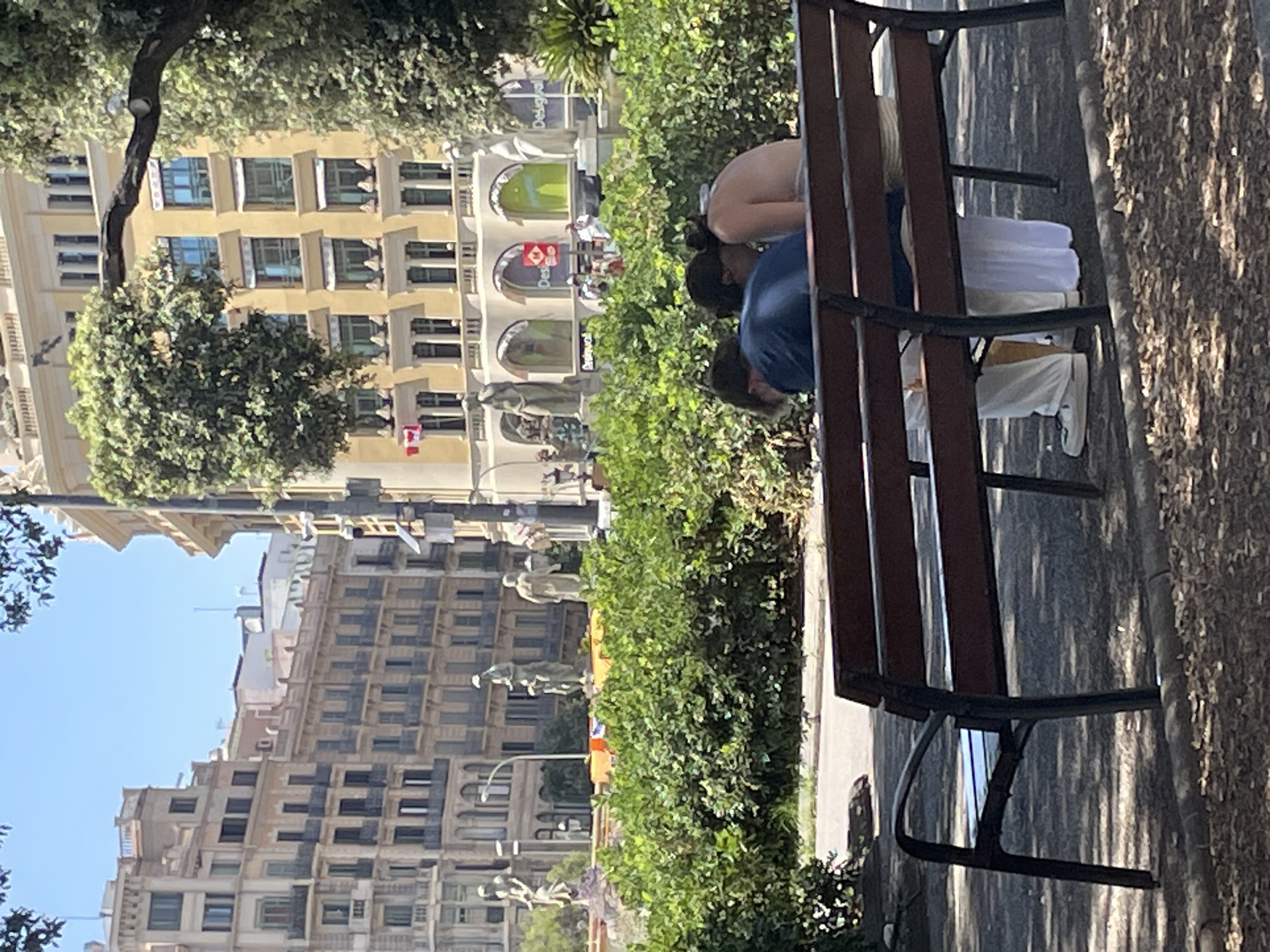 This is a vertical, medium-angle shot of a park bench and people in an urban setting. Three individuals are visible on the bench. Two are hunched over, one with their back to the camera, wearing a light-colored top and white pants, and the other in a blue shirt, also hunched over. The third person, with dark hair, is partially visible in the background, also in a light-colored top. The bench is made of dark wood slats and a dark metal frame.
The background shows a vibrant urban environment with buildings and greenery. To the left of the bench, there are lush green bushes and trees, partially obscuring the view of buildings. The buildings appear to be in a Mediterranean architectural style, with multiple stories, arched windows, and ornate details. One building has a yellow facade. There are also statues and streetlights visible in the mid-ground. The sky is a clear, bright blue, suggesting it's a sunny day, likely daytime given the lighting and shadows cast by the trees. The shadows on the ground suggest the sun is fairly high.
There is some visible text on the buildings in the background, including "DELGADO" which appears in a green archway. There is also a red square sign with white text and a Canadian flag. The overall atmosphere appears to be casual and relaxed, with people enjoying a day outdoors in a city park. The image is slightly blurry in some areas due to movement or focus.