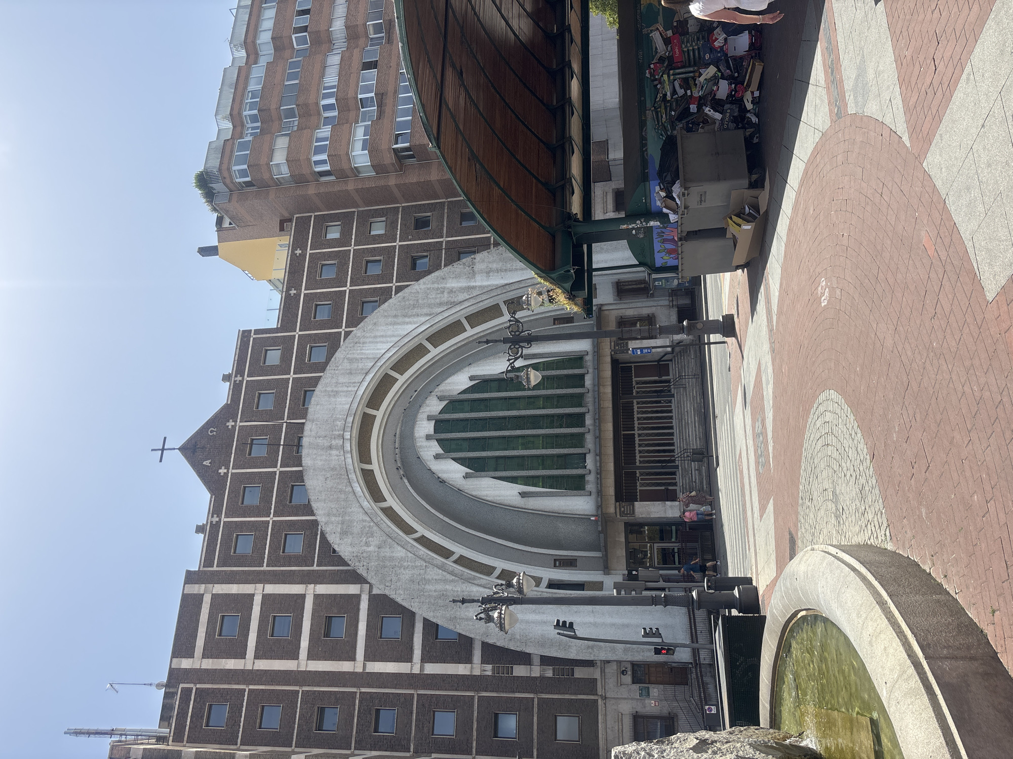 The image depicts a street scene in Valladolid, Spain, with a clear sky suggesting a sunny day. The orientation of the image is vertical, with the bottom of the frame showing a circular fountain filled with green water. Adjacent to the fountain is a paved area with a patterned design of reddish-brown bricks and light-colored stones.
Dominating the background is a large, imposing building with a curved, arched facade. This building appears to be a church or a similar religious structure, indicated by a cross at its apex and decorative elements resembling religious symbols. The building is constructed with dark bricks and features numerous small, square windows arranged in a grid pattern. Another taller, modern brick building with many windows rises behind it.
In the mid-ground, a covered market or a similar structure with a curved wooden roof is visible. In front of this structure, a pile of discarded items, including boxes and various objects, is accumulated, suggesting a public space that may also serve as a collection point. A few people are present in the scene, some sitting near the market area, while others are likely passing by. The overall impression is of an urban environment on a bright day, with a mix of historical and modern architecture.
