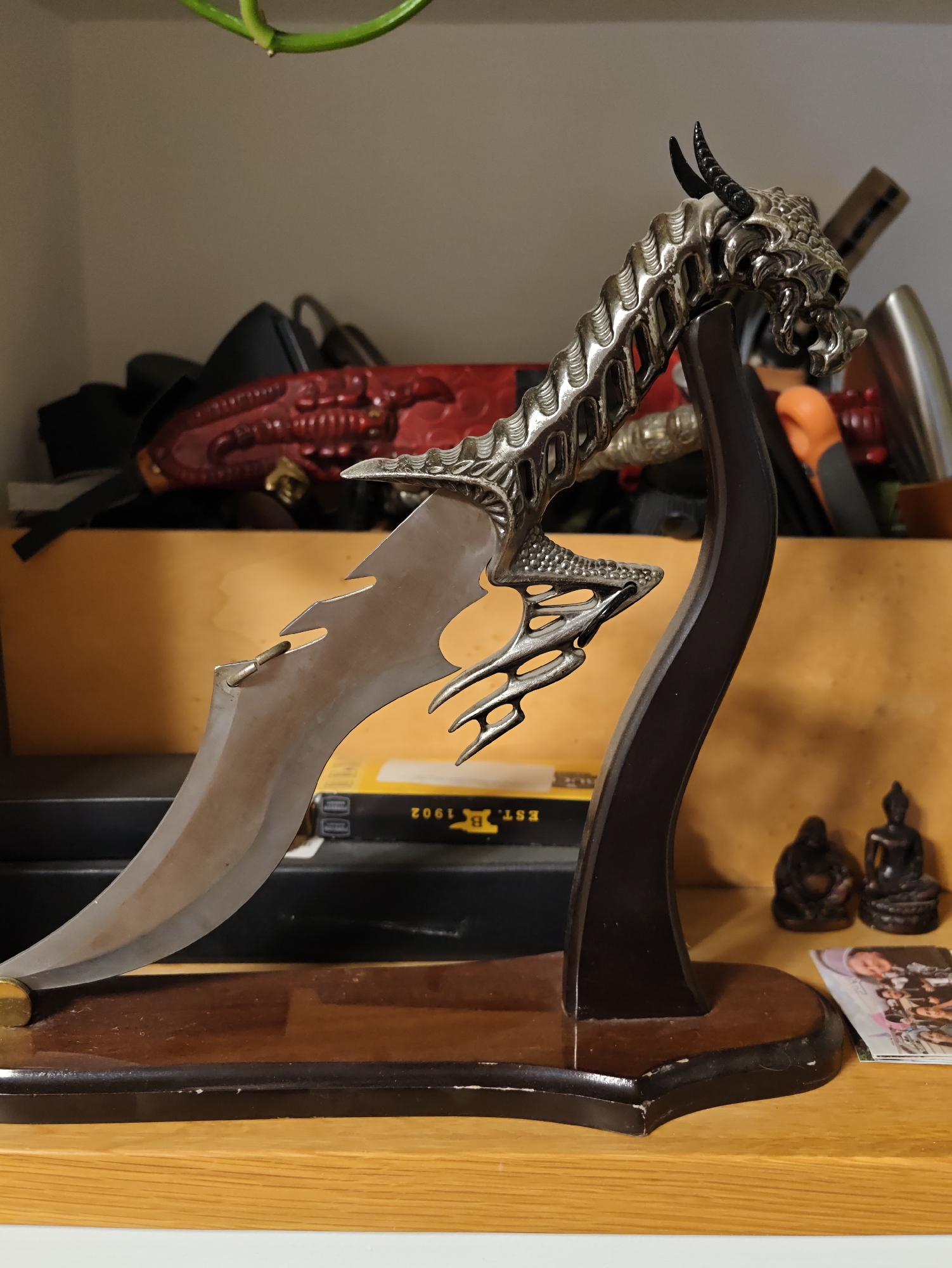 An ornate dragon-themed dagger is displayed on a wooden stand on a shelf. The dagger features a detailed silver dragon head as the pommel, with intricate scales and horns. The blade is curved and sharp, with a serrated edge near the hilt. The stand is made of dark wood with a polished finish. In the background, other objects are visible on the shelf, including what appears to be a red, textured item, possibly a scabbard or decorative piece, and other indistinct items. Two small Buddha figurines and a magazine or photograph are also present on the shelf to the right of the dagger. The setting appears to be indoors, on a wooden shelf. The location is not determinable from the image.