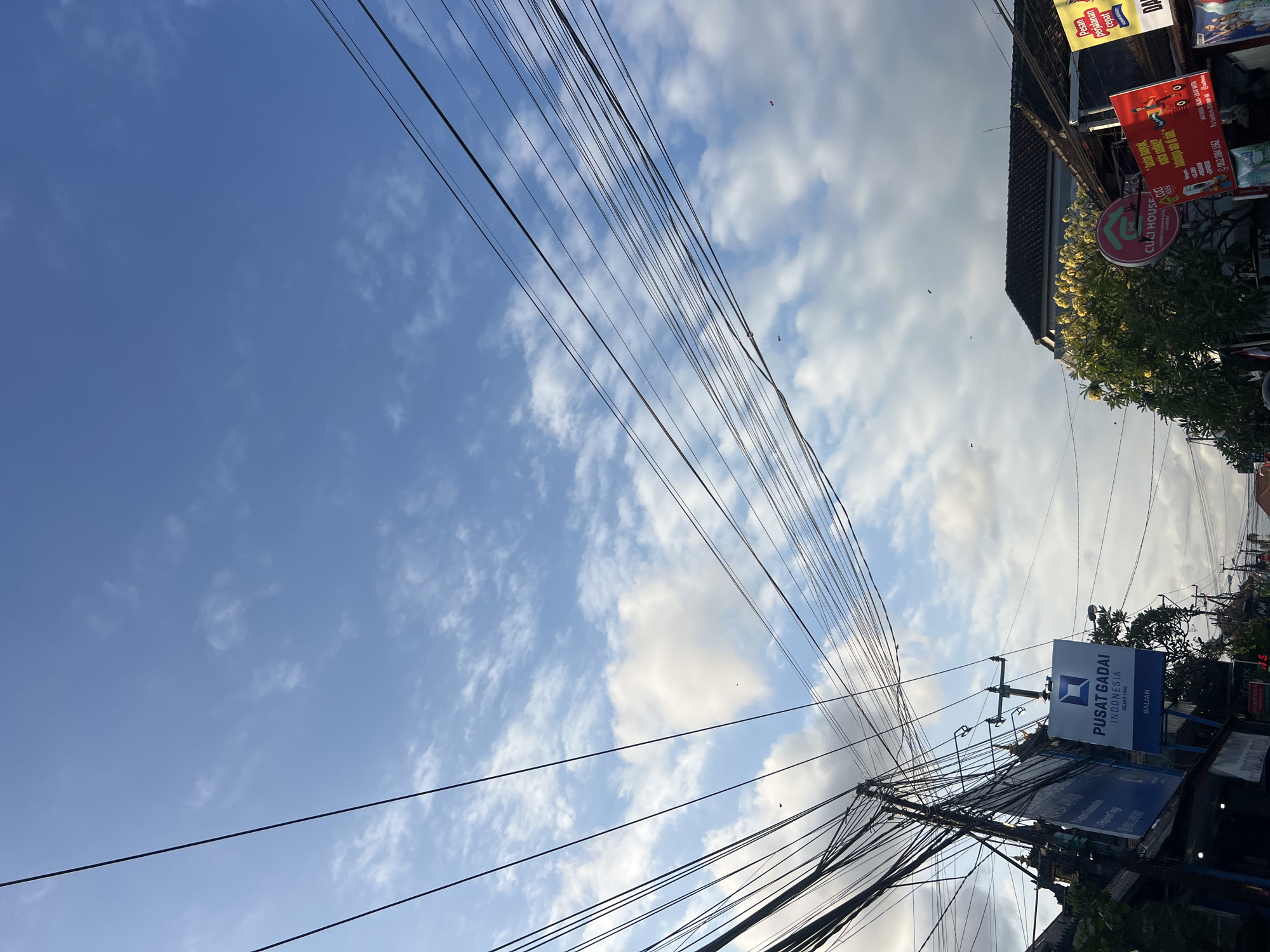 The image captures a street scene in Denpasar, Indonesia, characterized by a dense network of overhead utility wires against a partially cloudy blue sky. On the left, a sign reads "PUSAT GADAI INDONESIA SEJAK 1990 BALIAN." Further down the street, another sign, partially obscured, advertises "CUCI HOUSE." A tree with yellow flowers is visible on the right, along with storefronts. Several birds are seen in the sky. The overall impression is that of a typical urban environment in Denpasar with its characteristic overhead wiring.