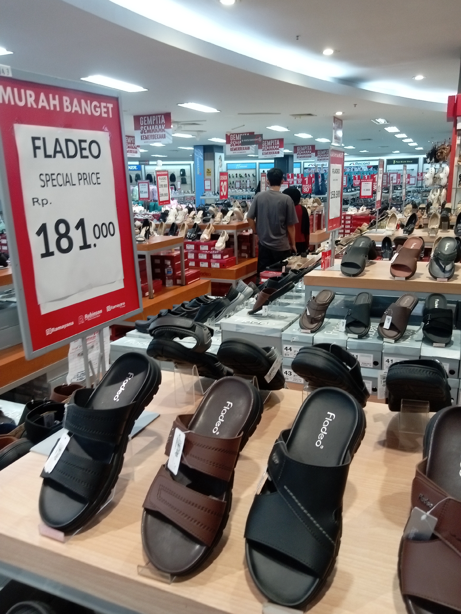 This is a medium shot, eye-level, indoor photograph of a shoe store display. The foreground features a selection of men's sandals on a wooden shelf. The sandals are mostly black and brown, with various strap designs. Some are on display stands, while others are placed directly on the shelf. In the background, more shoes and footwear are visible on shelves and in boxes. A large red sign on the left side of the image advertises "FLADEO SPECIAL PRICE Rp. 181.000" in large white lettering. Several overhead fluorescent lights illuminate the store. Two men are partially visible in the mid-ground, walking away from the camera. One man wears a grey t-shirt and the other wears a dark red shirt. The overall impression is a retail environment showcasing footwear. Based on the signage and common retail chains in Indonesia, this scene is likely within a department store or a large shoe retailer in Batam, Indonesia, possibly Robinsons or Ramajaya.