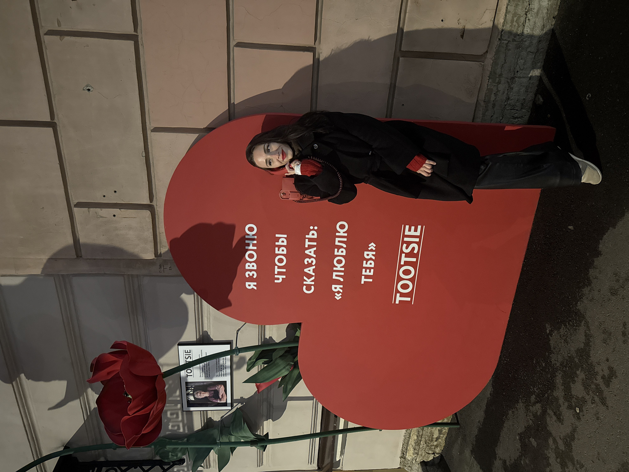 A sideways, full shot depicts a dark-haired woman in a dark coat with red sleeves lying on a large, red heart-shaped sign. The sign has Cyrillic text which translates to "I call to say 'I love you'" with "TOOTSIE" written below in English. The woman is holding a red telephone receiver to her ear.
The scene is an outdoor location, with the heart-shaped sign positioned against the side of a building with a textured, light-colored wall. A large artificial red flower on a green stem stands near the base of the heart. A sign with a picture of a person is next to the flower. The ground is paved.
The location is the Italian Garden in Saint Petersburg, Russia, as evidenced by the presence of the "Tootsie" installation, a public art piece.