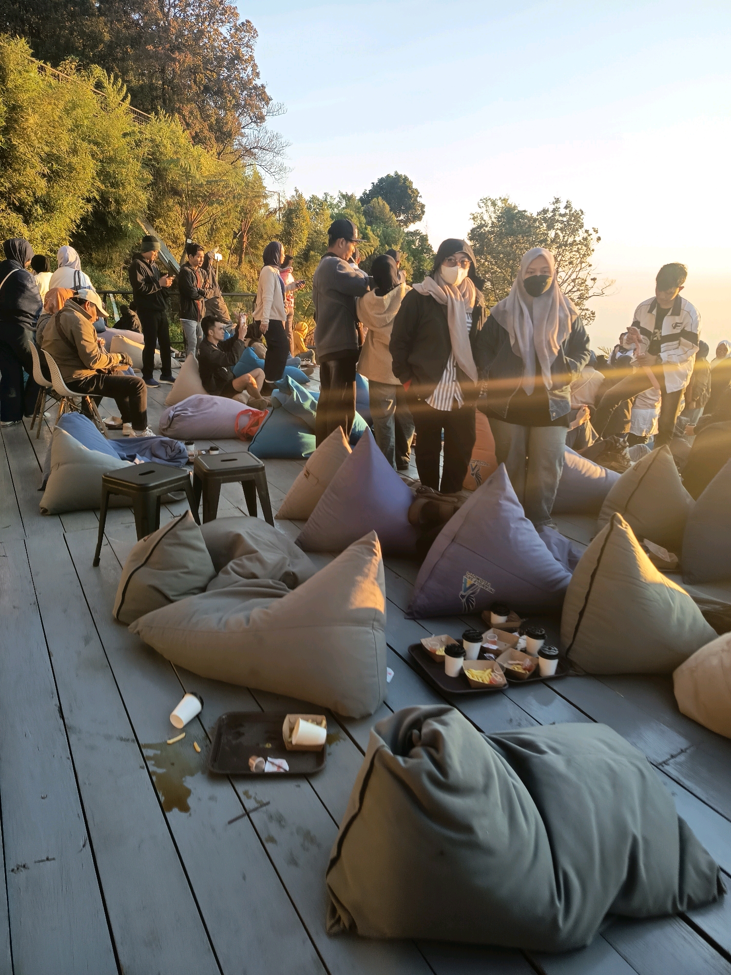 The image depicts an outdoor gathering on a wooden deck, likely in Keditan, Indonesia, during a sunset or sunrise given the warm lighting. A group of people, some wearing masks and headscarves, are casually seated on beanbag chairs and some chairs. In the foreground, several large beanbag chairs are arranged on the deck. A dark tray with food containers and cups is placed on the deck, with some food and liquid spilled nearby. In the background, more people are visible, some standing and interacting, while others are seated. Trees and foliage form a backdrop, with a hazy sky suggesting a mountainous or elevated location. The overall atmosphere appears relaxed and social.