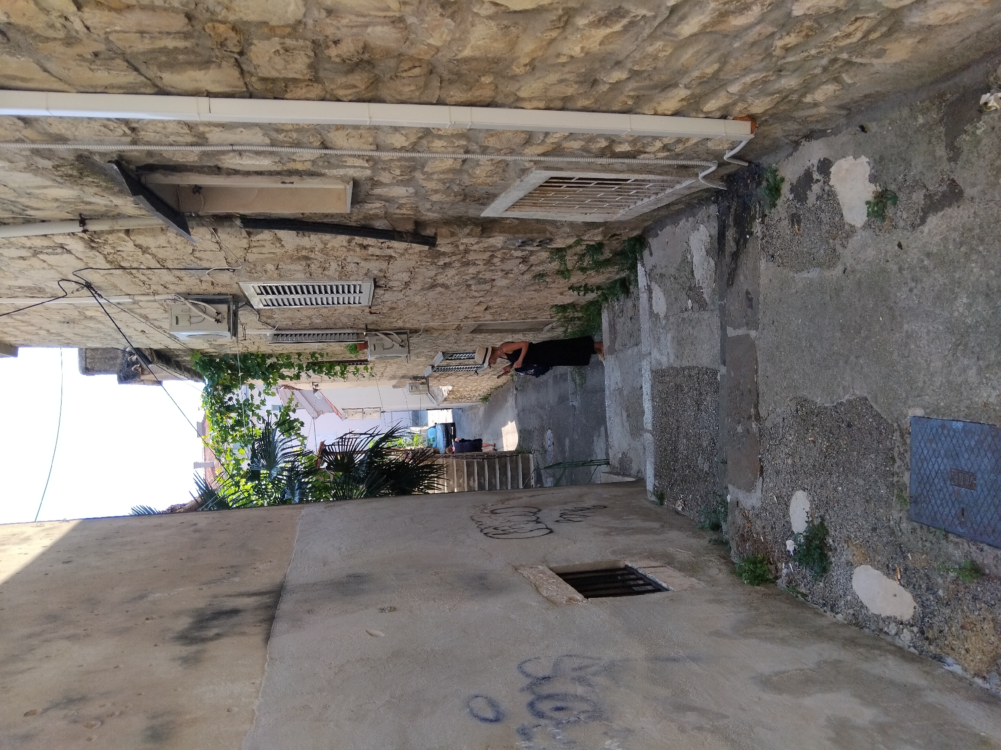 A narrow, stone-paved alleyway in Split, Croatia, slopes downwards between two buildings. The building on the left has a rough, light-colored plaster wall with graffiti. The building on the right is constructed of light brown stone blocks and features several windows, some with shutters, and air conditioning units. A woman in a dark dress and a straw hat stands on the stairs in the middle of the alley, looking at her phone. Further down the alley, another person is walking away, and a car is visible. Green foliage, likely palm trees and vines, spills over the walls from above. The ground is a mix of stone steps, gravel, and concrete.
