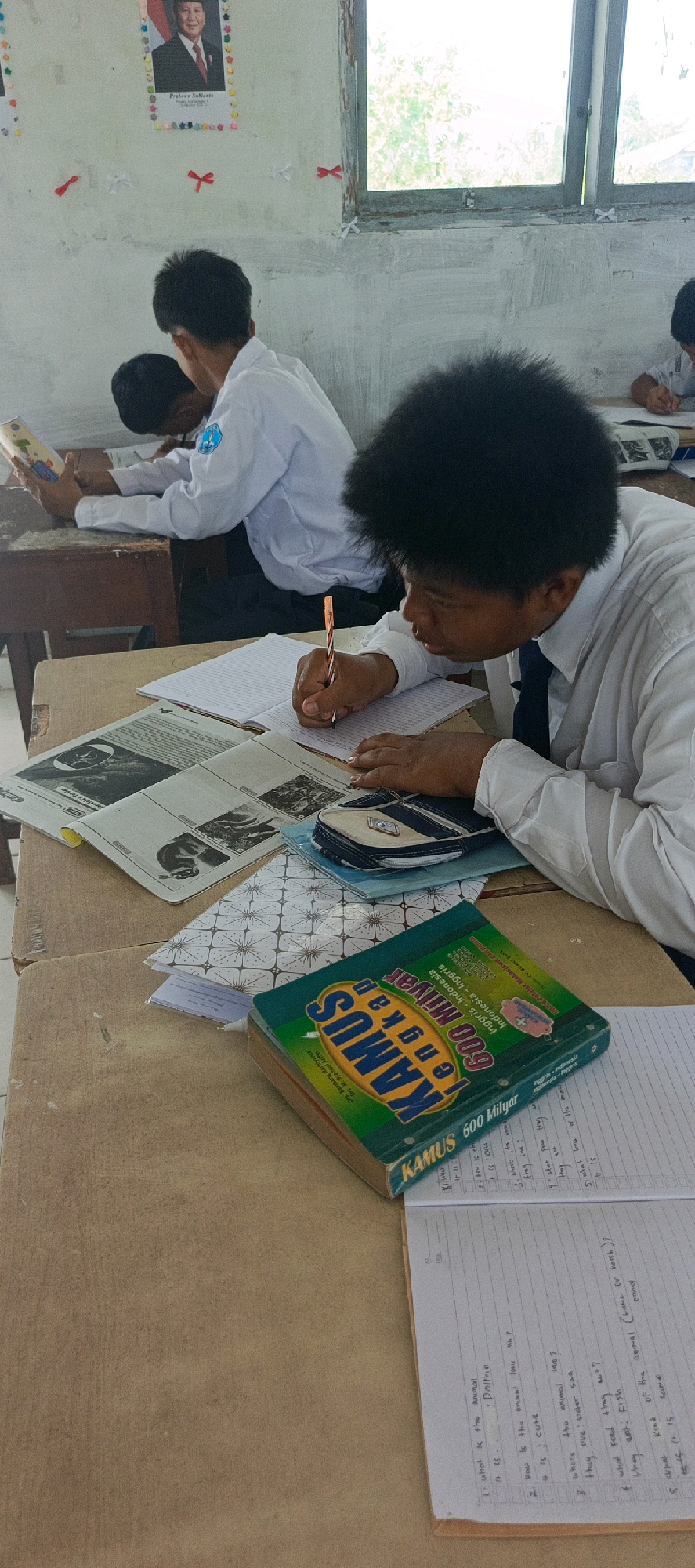 This image depicts a classroom scene in Desa Sarang Mandi, Indonesia, where students are engaged in learning. In the foreground, a young boy with voluminous black hair, wearing a white school uniform with a dark tie, is diligently writing in a notebook with an orange pen. His desk is covered with various educational materials, including a large green dictionary titled "KAMUS," an open newspaper with black and white images, and a small blue and white pencil case. Behind him, another student in a similar uniform is seated and appears to be reading a book. The classroom environment is suggested by the presence of a framed portrait of an Indonesian official on the wall and a window looking out onto greenery. The desks appear to be made of wood, and the overall impression is one of a typical educational setting in a rural Indonesian village.