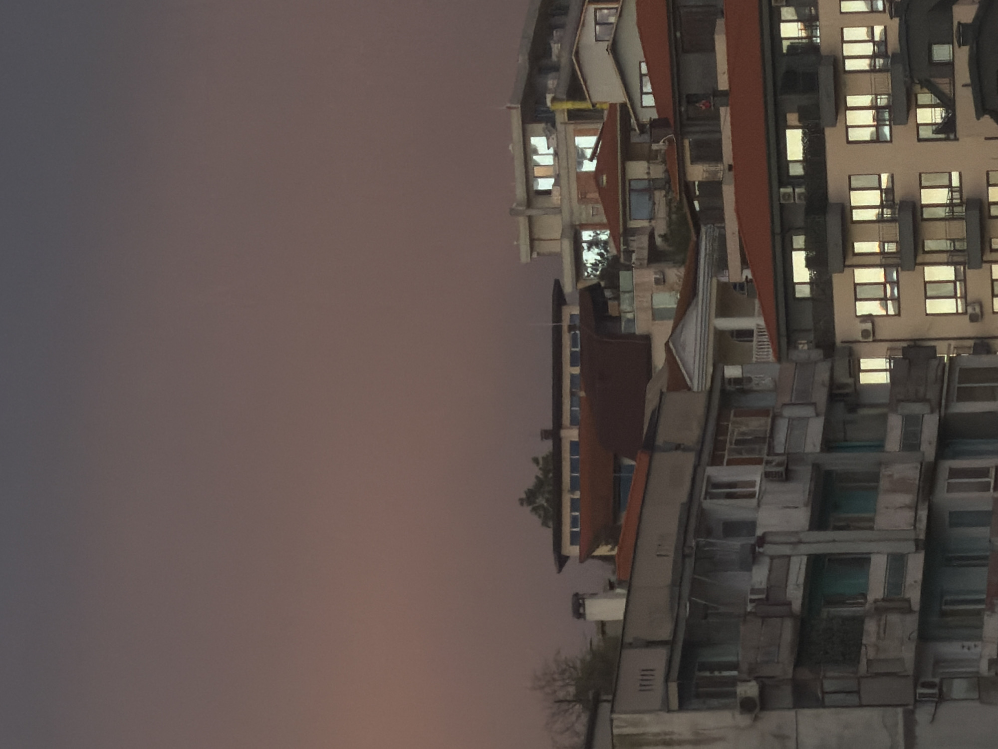 A vertically oriented, low-angle shot depicts a cluster of buildings against a dusk sky. The buildings are varied in design and height, featuring a mix of architectural styles. Some have red-tiled roofs, others are flat. Many have windows, some with lights visible inside. Air conditioning units are visible on some buildings. The sky is a gradient of purple, grey, and orange, suggesting twilight. A leafless tree stands in front of the buildings. There is no evidence to determine the location within the city of Varna, Bulgaria.