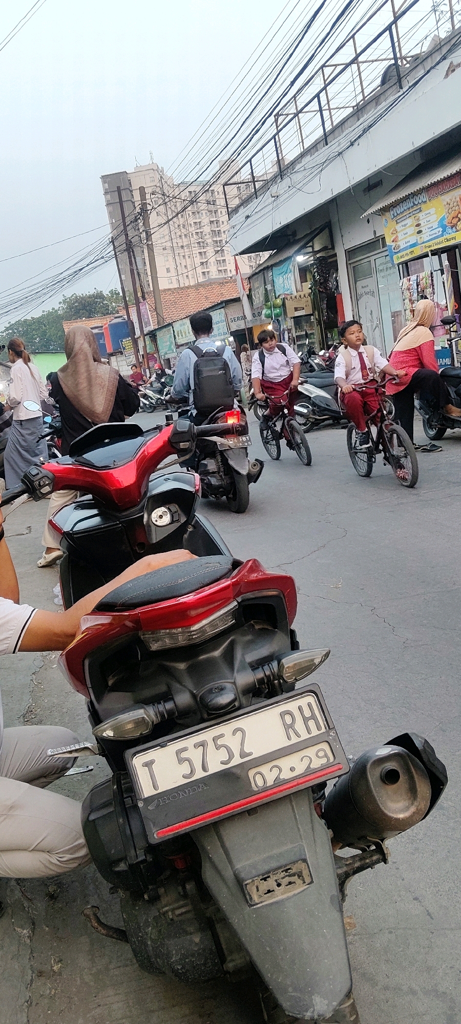 In this medium shot, two young boys in school uniforms, a white shirt and red pants, ride bicycles down a busy street in Mekarmukti, Indonesia. They are accompanied by several adults on motorcycles, also in the street. The scene appears to be during the day, with a bright, overcast sky. The street is lined with shops and buildings, and there are many power lines overhead, suggesting an urban or semi-urban environment. The buildings appear to be made of concrete and brick, with some shops having awnings and signs. One sign visible is for "Frozen Food DUO SLALLABA" and another that reads "SERBA 0.00". A red Honda motorcycle is prominently featured in the foreground, with its license plate reading "T 5752 RH" and "02.29". The overall atmosphere is one of everyday activity and local commerce.