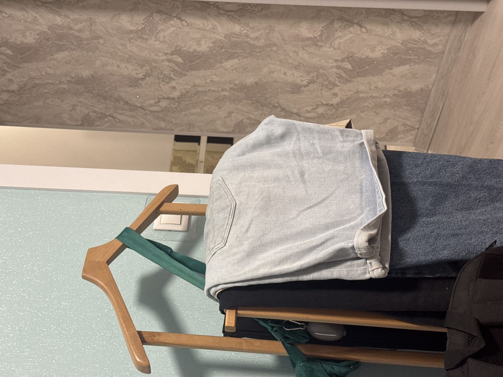 A stack of folded clothing rests on a wooden clothes hanger. The clothing consists of light blue denim jeans on top of black fabric, with darker blue denim partially visible beneath. A green garment and a black backpack are also present in the composition. The hanger is positioned against a light blue wall, and a white electrical outlet is visible in the background. Above the wall, the ceiling has a marbled pattern in shades of gray and white. The location is not indicated within the image, and thus, Izhevsk, Russia cannot be confirmed.