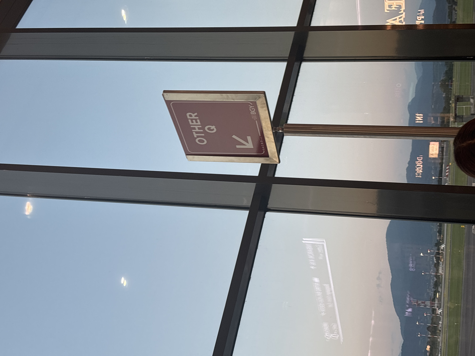 The image captures a view through a large window, likely at Orio al Serio International Airport in Bergamo, Italy. In the foreground, a maroon sign with a silver border is mounted on a metal pole. The sign displays "OTHER Q" in white text, with a white arrow pointing left below the letter Q. At the bottom of the sign, "SACRO..." and "BGY" are visible in smaller text, with "BGY" likely representing the IATA code for Bergamo. The window reflects the sky and distant mountains, as well as signs inside the terminal. Through the window, an airport tarmac is visible with a control tower and various airport lights, along with a range of hills or mountains in the background. The sky is clear and appears to be either early morning or late evening, judging by the soft, diffused light. The overall scene depicts an airport environment with a focus on wayfinding signage.
