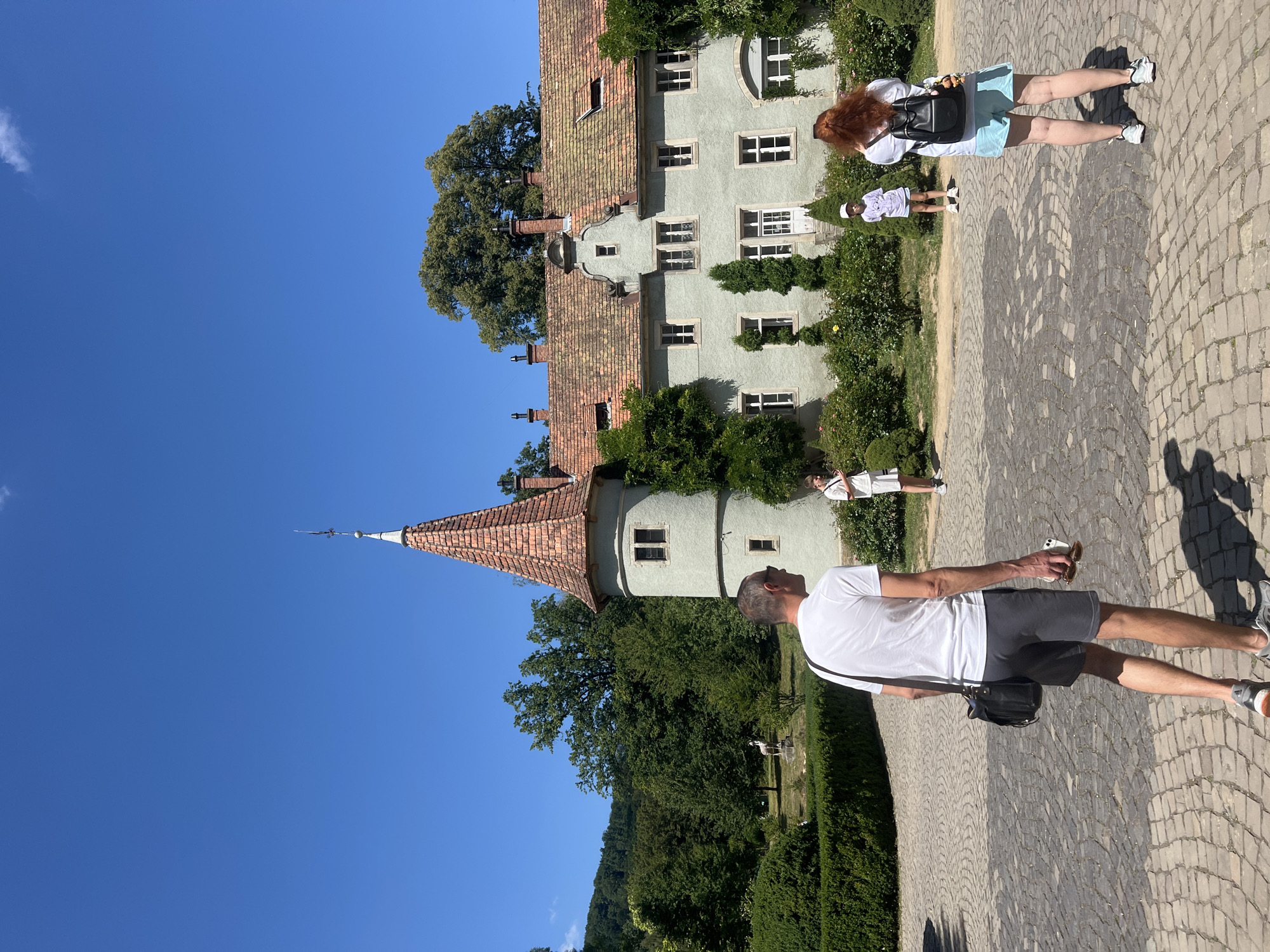 In a courtyard paved with cobblestones, a light green building with a red tiled roof stands under a clear blue sky. The building has a tower with a pointed roof, multiple windows, and chimneys. It is surrounded by lush green trees and bushes, including manicured hedges and rose bushes. In the background, a small statue of a deer is visible.
Four people are in the courtyard. In the foreground, a man with a white t-shirt and gray shorts walks away from the camera, carrying a black shoulder bag and a mobile phone. To his right, a woman with red hair, wearing a white t-shirt and blue shorts, is facing away from the camera, carrying a black backpack. Further in the background, two children, a boy and a girl, stand near the building. The boy wears a white t-shirt and shorts, while the girl wears a light purple t-shirt and shorts. They appear to be looking at the building. The scene is set in the city of Karpaty, Ukraine.
