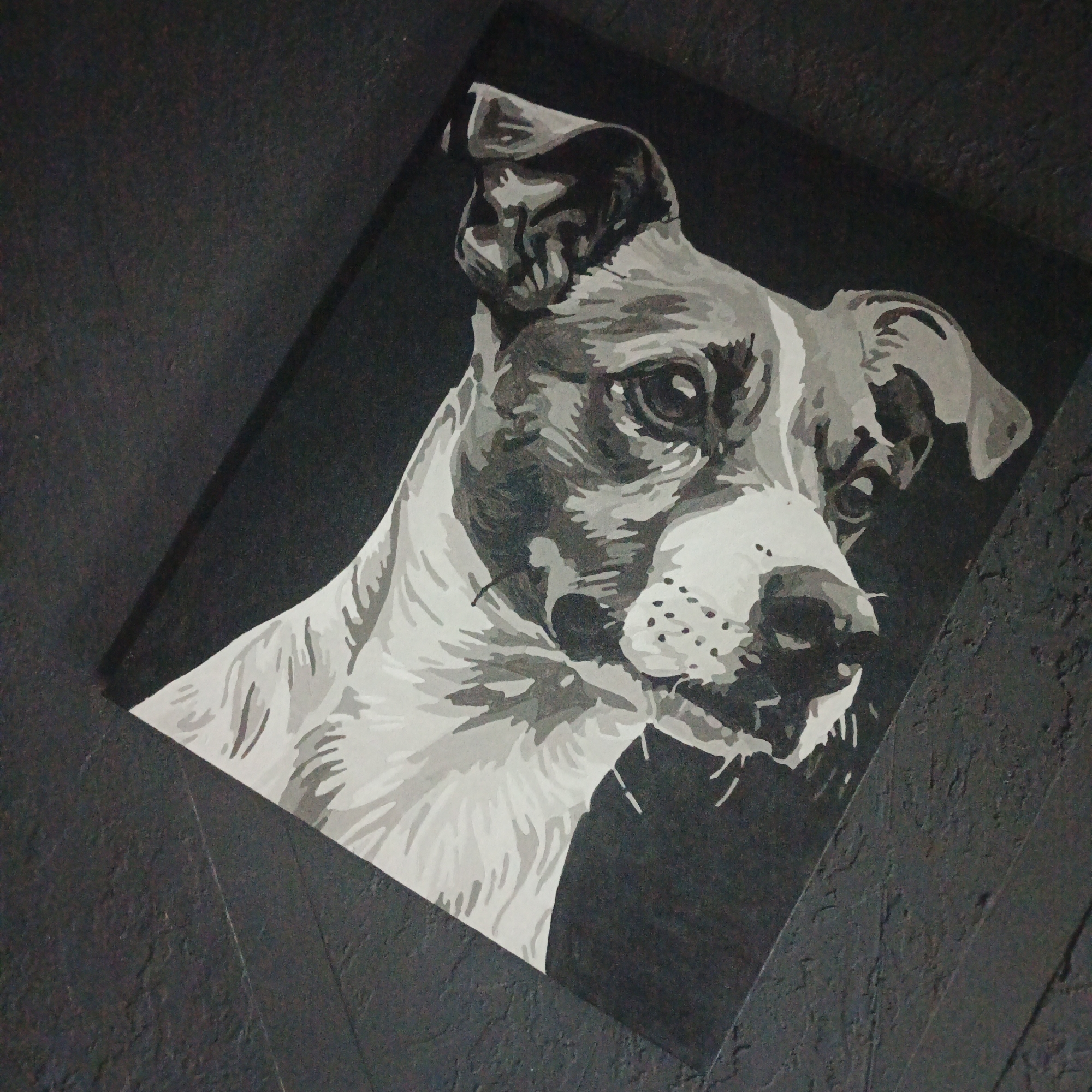 The image displays a painting of a dog's head, rendered in grayscale. The dog is positioned on a dark, textured surface that appears to be a wall. The painting itself is tilted at an angle, adding to the dynamic composition. The dog's face is detailed, with expressive eyes and a discernible snout. The fur is depicted with varying shades of gray, creating a sense of depth and texture. The background of the painting is solid black, which makes the dog's features stand out. The overall impression is that of a striking portrait. The location of this painting in Vyshgorodok, Russia, is not visually evident from the image itself.