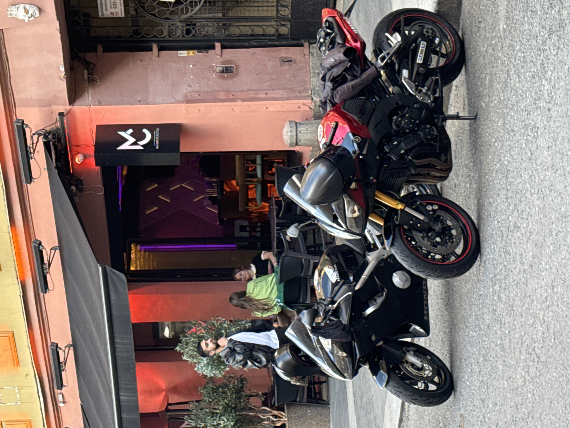 This is a medium shot of a street scene in Saint Petersburg, Russia, featuring two motorcycles parked in front of a building with an outdoor seating area. The building appears to be a cafe or restaurant, with a black awning and a pink facade. A sign with a stylized "MC" logo and text in Cyrillic is visible. In the background, two women are seated at a table, and a man in a black leather jacket and white t-shirt is standing and looking to his left, with his hand near his chin. The motorcycles are the primary subjects in the foreground, a black Honda on the left and a red Yamaha on the right. Both are sport bikes. The pavement is asphalt, and a stone curb is visible. The overall impression is an urban street scene during daylight.