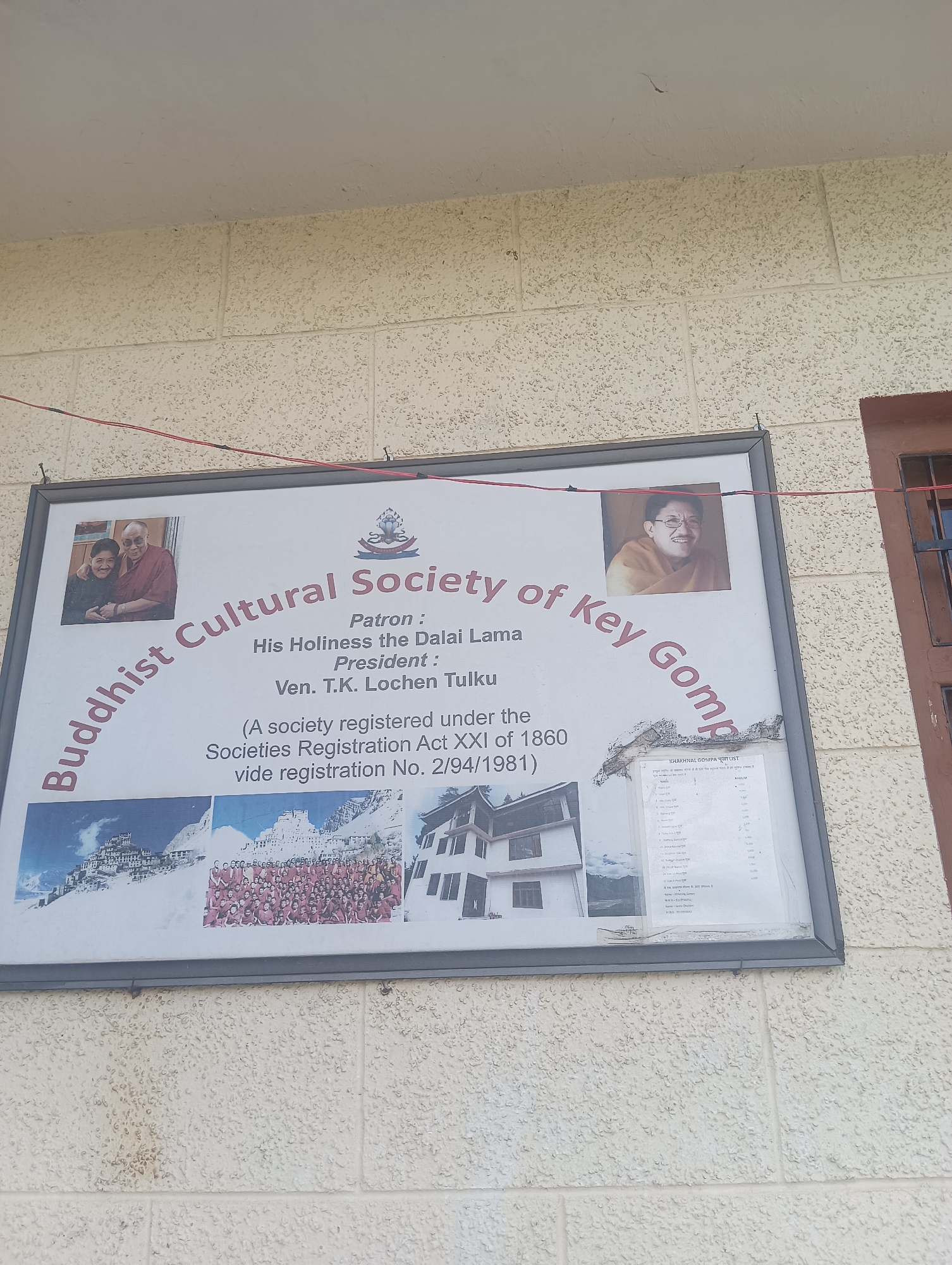 The image shows a framed sign mounted on a light-colored, textured brick wall. The sign displays text and photographs. Dominating the left side of the sign is the curved text "Buddhist Cultural Society of Key Gomp". To the right of this text is a crest, and below that, text indicating "Patron: His Holiness the Dalai Lama" and "President: Ven. T.K. Lochen Tulku". Further down, it states "(A society registered under the Societies Registration Act XXI of 1860 vide registration No. 2/94/1981)".
The top left of the sign features a photograph of two men, one in a maroon robe and the other in a darker robe. The top right of the sign has a portrait of an older man wearing glasses and an orange robe. Below the text, there are several smaller photographs depicting buildings and a large group of people in maroon robes. In the bottom right corner of the sign, there is a list in a smaller font, potentially detailing donations or members, with a heading that appears to be "KHAKHNAL GOMPA PUJA LIST". The overall sign is framed by a dark border and is slightly weathered at the bottom. The brick wall has some staining and discoloration. A red wire is visible running horizontally above the sign, attached to the wall by hooks. A portion of a dark-colored window frame with bars is visible on the right edge of the image. The scene appears to be outdoors, likely on the exterior of a building.