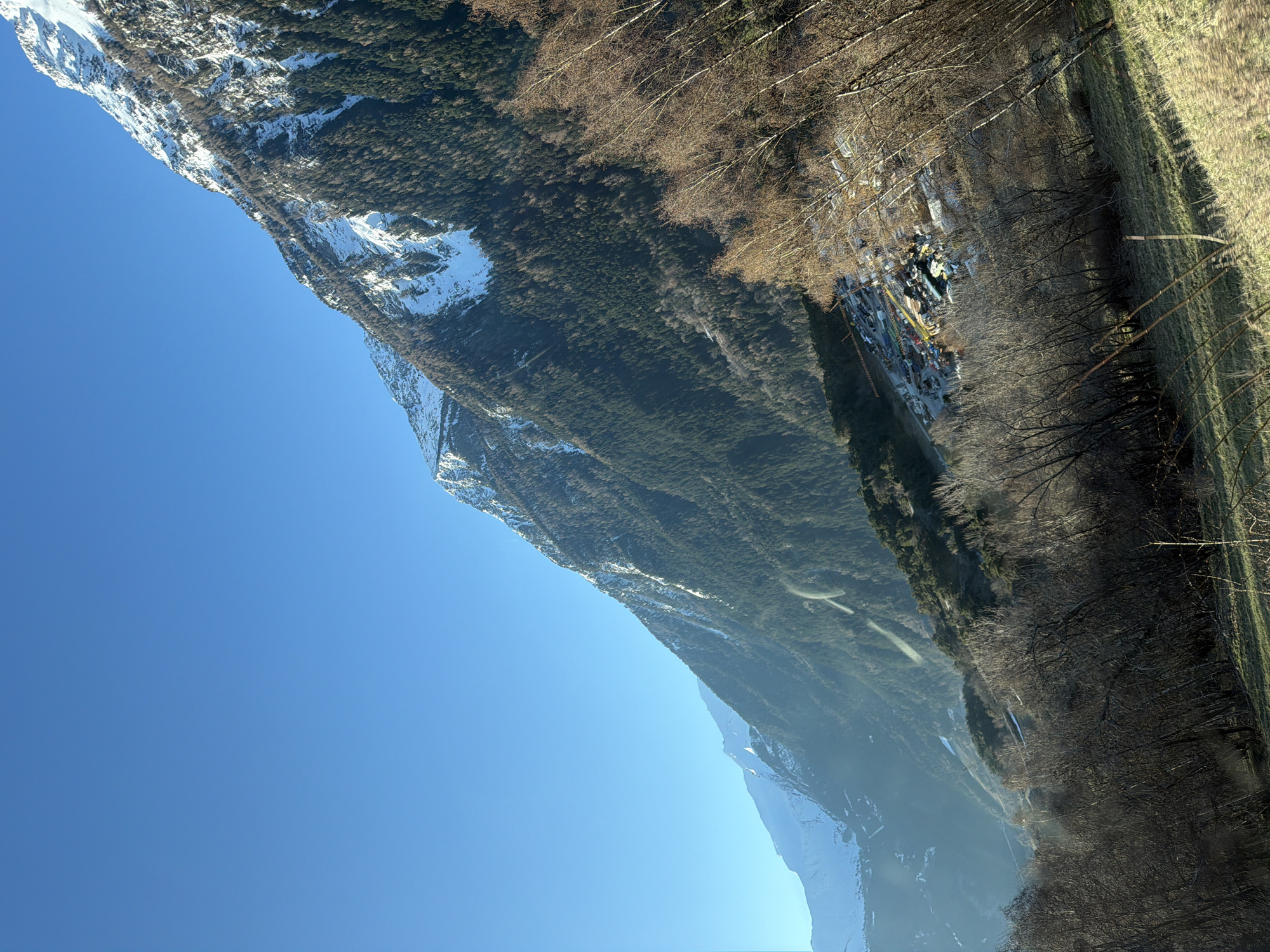 The image depicts a mountainous landscape on a clear, sunny day. The sky is a bright, cloudless blue. The left side of the frame is dominated by a mountain range with patches of snow at higher elevations. A dense evergreen forest covers the lower slopes of the mountains. Along the base of the mountains, where the forest meets the valley floor, is a narrow strip of buildings, likely a settlement or small town, which can be identified as part of Saint-André, France. To the right of the forest, the landscape transitions to more open areas with bare trees and grassy fields.