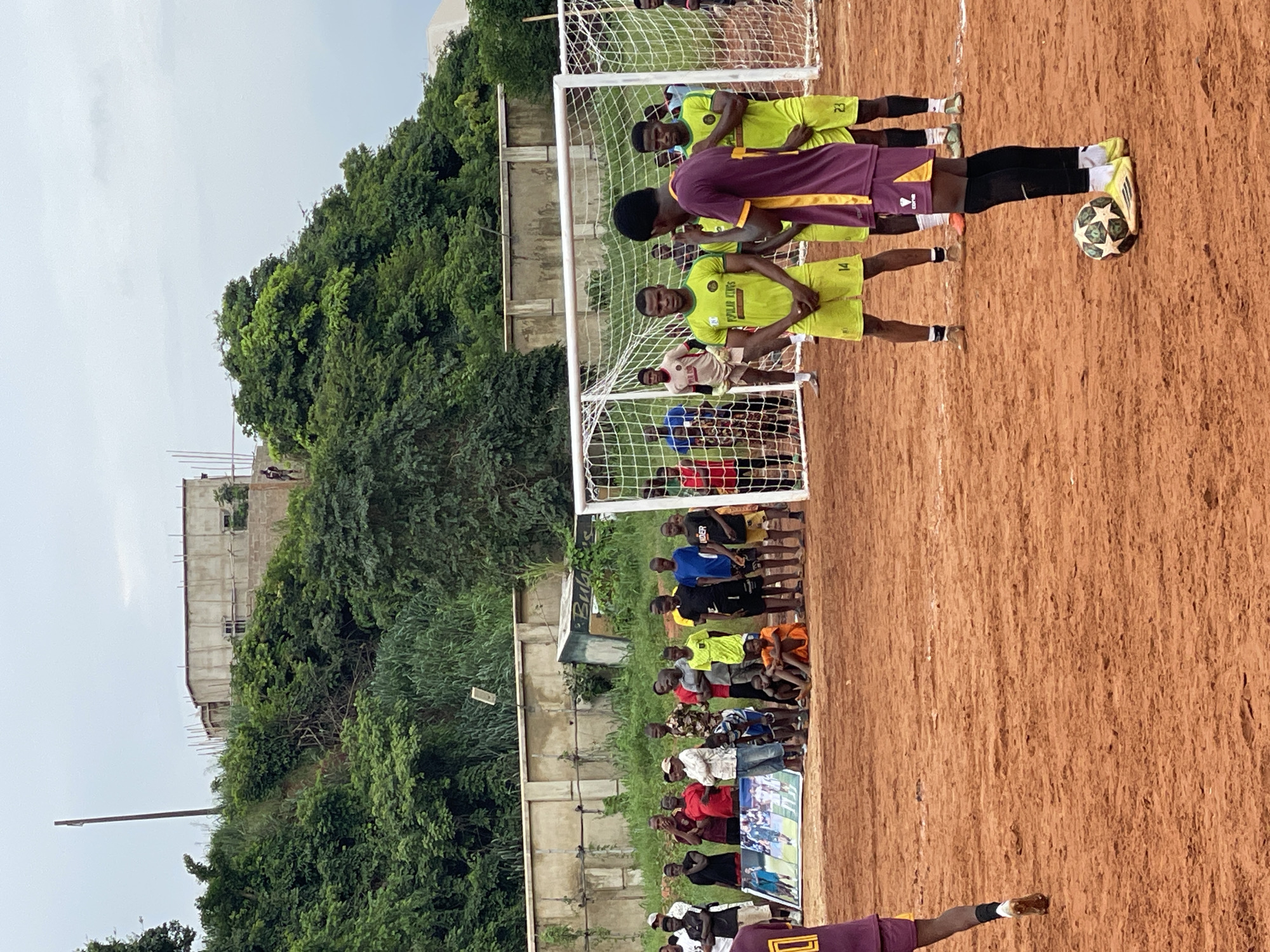 In an outdoor setting with a dirt field and a cloudy sky, a group of young men and boys are gathered, seemingly for a soccer match. A soccer goal with a white net is visible in the background. Several players in athletic wear, primarily lime green jerseys and shorts, stand on the field. Some are in various poses, looking towards the goal or engaging with each other. A soccer ball rests on the ground near the feet of one player. Spectators, dressed in casual clothing, stand behind a low wall, observing the scene. In the far background, a partially constructed building is visible on a grassy hill, with trees and vegetation surrounding it. The overall impression is that of a local sporting event taking place in a semi-urban or rural environment.