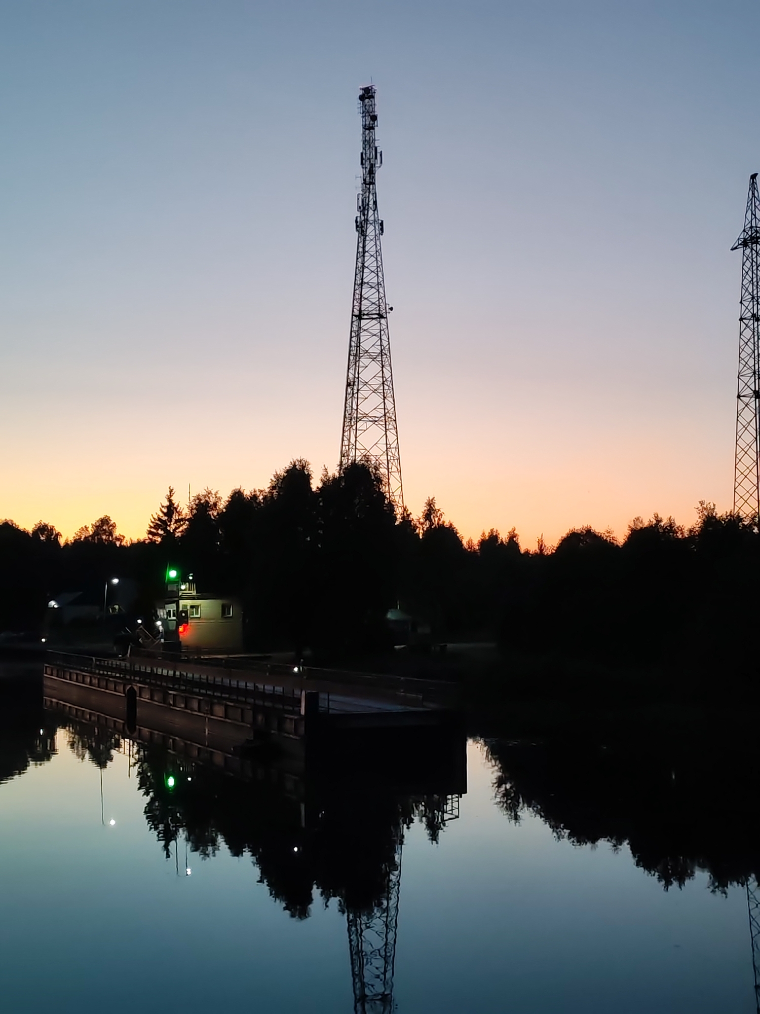 At dusk in Annensky Most, Russia, a body of water reflects the soft hues of the sky and the dark silhouettes of trees. On the far bank, a small building with green and red lights is visible, situated amongst the trees. A tall, lattice-like communication tower rises into the sky behind the building, and another similar tower is partially visible on the right side of the frame. A man-made structure, possibly a dock or a bridge, spans the water in the foreground, also reflected in the calm surface. The scene is characterized by a serene, twilight atmosphere, with the towers indicating a connection to infrastructure within the otherwise natural landscape.