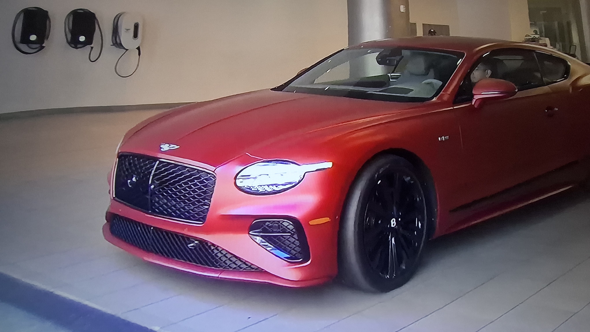 A red Bentley Continental GT coupe is parked indoors. Three charging stations are mounted on the wall to the left of the vehicle. A person is seated in the driver's seat. The location is Unknown, Unknown.