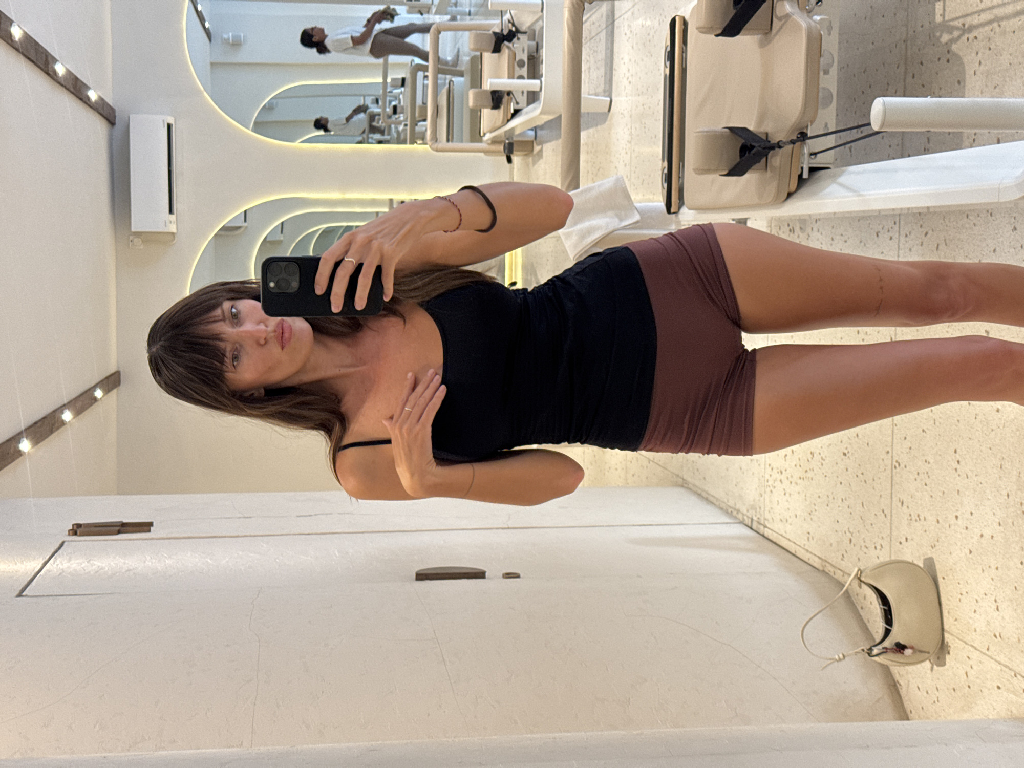 A woman is taking a mirror selfie in a room, possibly in Cemagi, Indonesia, given the prompt. She is wearing a black tank top and reddish-brown shorts. In the background, several white exercise machines are visible, along with a large decorative mirror with organic-shaped cutouts. A cream-colored purse is on the floor next to the wall. The room appears to be a fitness or Pilates studio. The wall and floor are off-white with a speckled pattern.