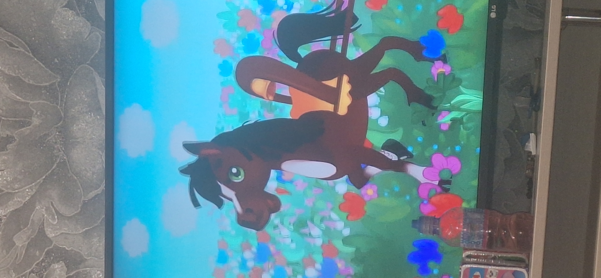 A cartoon horse stands in a field of flowers. The horse is brown with a black mane and tail, and it is wearing an orange saddle. The horse has its mouth open and appears to be smiling. The flowers in the field are various colors, including pink, red, blue, and yellow. The sky is blue with a few white clouds. The image is a still from a video, as evidenced by the television screen it is displayed on, and the shelf beneath it. There are several objects on the shelf, including a bottle of liquid and what appears to be a phone case. The image appears to be taken indoors, and the wall behind the television is textured. The location of this scene in Bila Tserkva, Ukraine cannot be determined from the image alone.