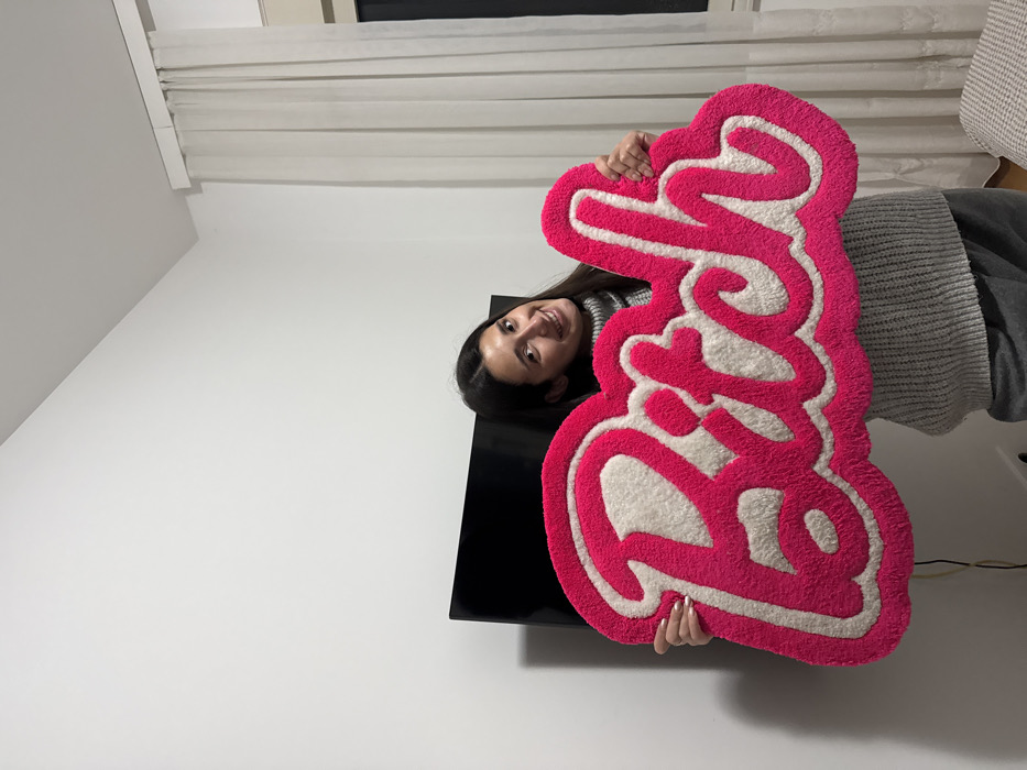 A smiling woman holds a pink and white custom rug that says "Barbie" in cursive font. She is wearing a gray sweater and standing in a room with white walls and light-colored curtains. The rug is large and fluffy. A black television is seen behind the rug.