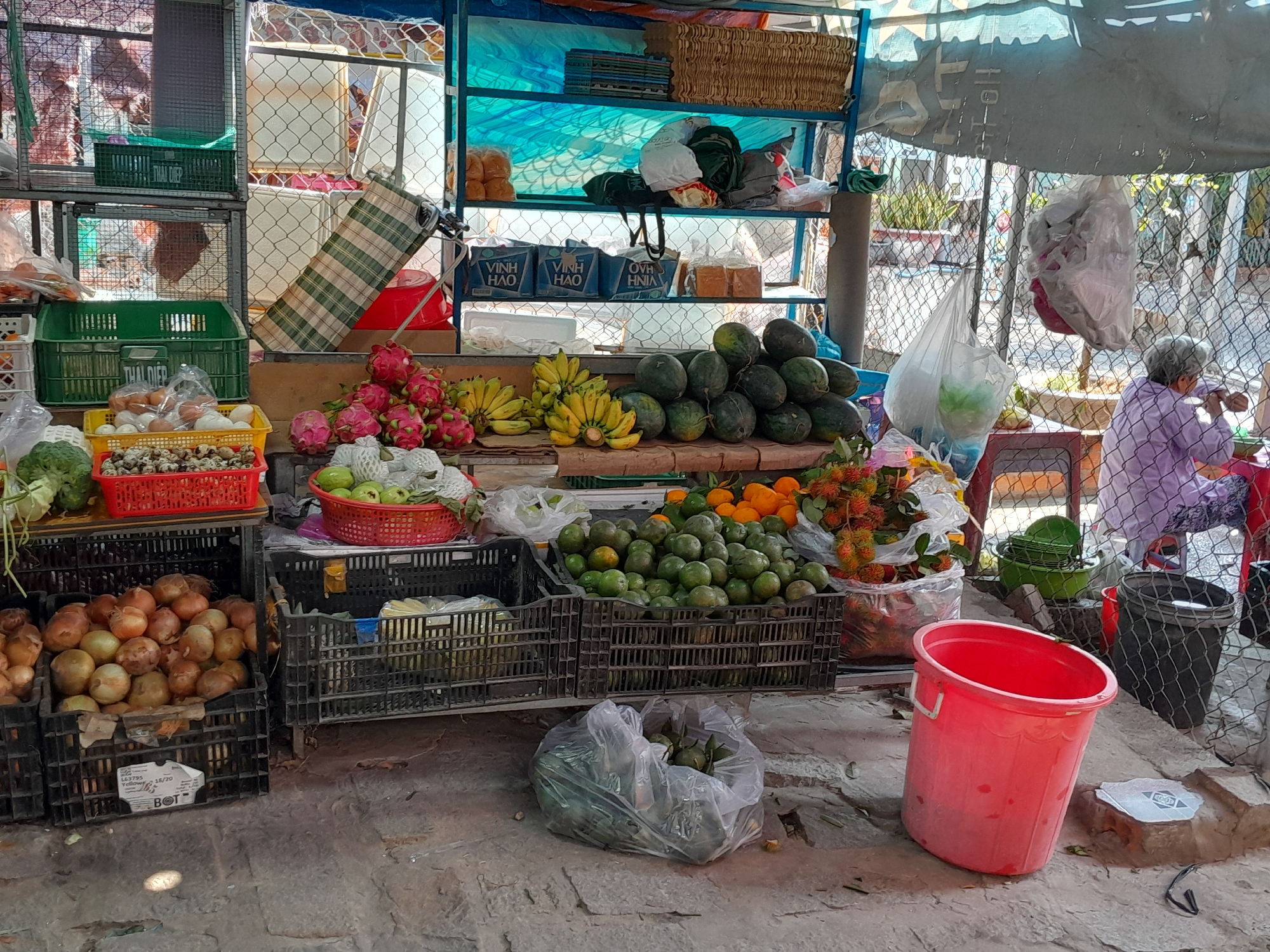 This is an outdoor scene at a market in Phu Thuy Ward, Vietnam, featuring a stall laden with fruits and vegetables. A woman, partially obscured by a chain-link fence and a tarp, is seated to the right, appearing to be engaged in an activity. The stall displays various items including dragon fruit, bananas, watermelons, oranges, and onions arranged in crates. Several plastic baskets and buckets, including a prominent red one in the foreground, are also visible. The ground is paved with rough concrete. The overall atmosphere suggests a typical market environment with goods for sale and people going about their day.