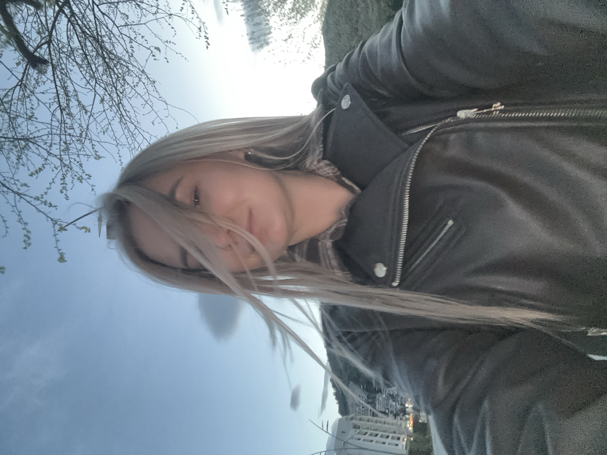 A young woman with light blonde hair, wearing a black leather jacket over a patterned sweater or top, appears to be taking a selfie outdoors. The background is a blurred cityscape of what appears to be the city of Budva, Montenegro. In the background, buildings can be seen; the cityscape is reflected on the water. Bare tree branches are also visible. The sky is overcast or hazy. The photo seems to be taken at an oblique angle.