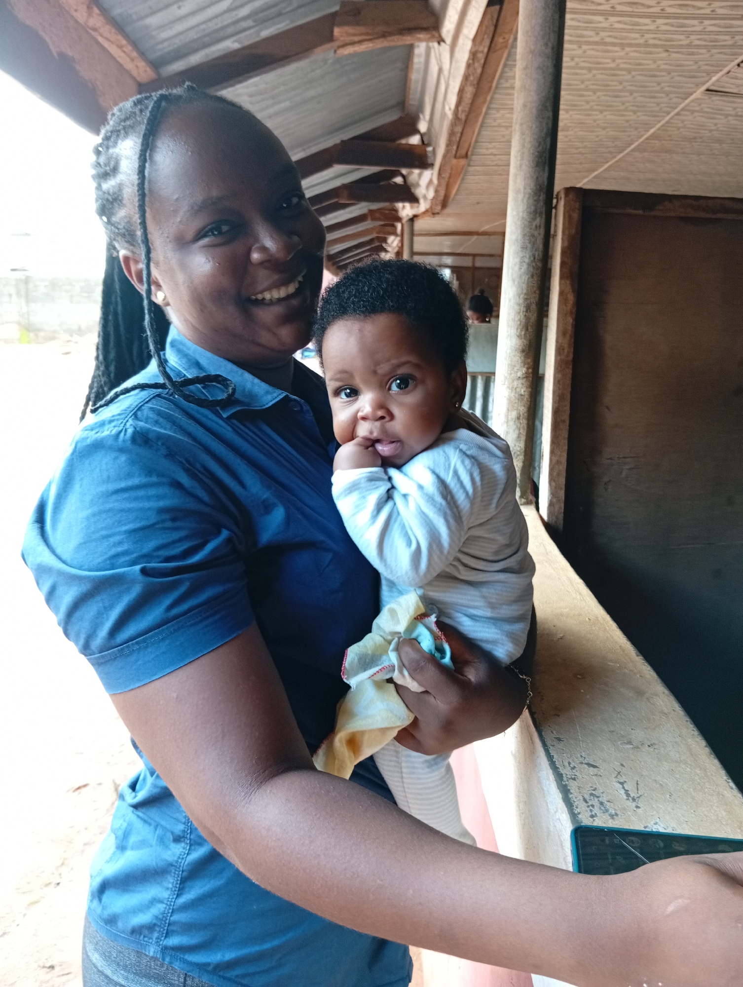 A woman is holding a baby in what appears to be a covered or semi-enclosed outdoor space. The woman is smiling at the camera, and she has dark skin, braided hair, and is wearing a blue button-down shirt. The baby, also with dark skin, is looking directly at the camera with a finger in its mouth. The baby is wearing a striped onesie and holding a small cloth. The location is Unknown, Unknown.
The background includes a ceiling with wooden beams and a wall. A vertical post is visible. A mobile phone is visible on the bottom right corner of the image. The scene is lit by natural light.