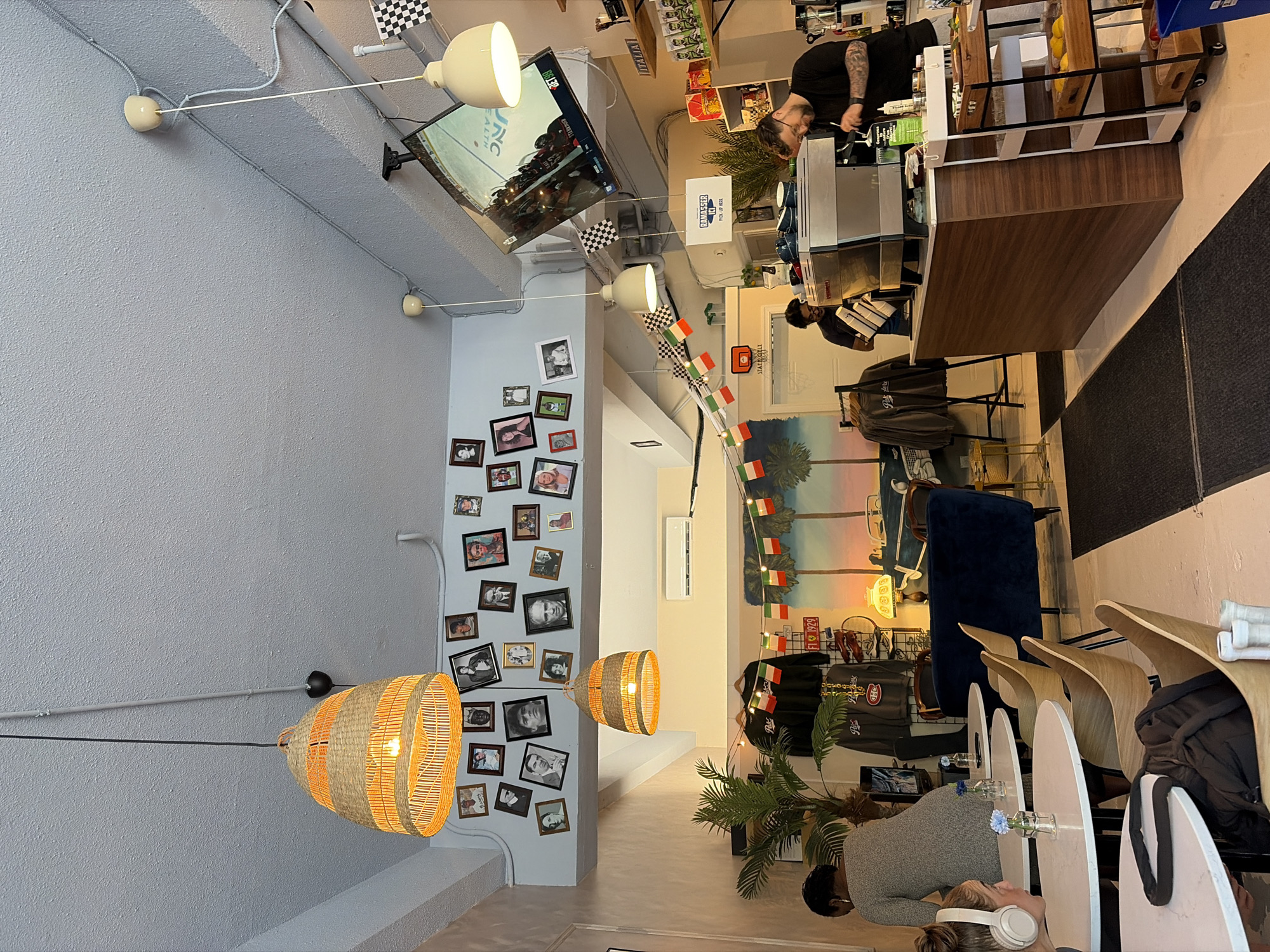 The image captures a cafe or restaurant setting, likely located in Montreal, Canada, indicated by the "RAMASSER ICI PICK-UP HERE" sign. The scene appears to be a hip, trendy space with unique design elements.
The interior is lit by pendant lights and spotlight fixtures. A television screen displays a hockey game, possibly an NHL match. There are checkered and Italian flags decorating the area, adding a touch of international flair.
A person wearing a black shirt and sporting arm tattoos stands behind the counter, possibly a barista preparing drinks. In the lower part of the image, individuals sit at tables, presumably customers engaging in conversation or enjoying their orders.
The walls are adorned with framed photographs, and there's a mural depicting palm trees and a sunset in the background, creating a tropical ambiance. Clothing with logos resembling the Montreal Canadiens (NHL team) is hanging on a display.
Furniture includes round tables with modern chairs, along with a blue upholstered chair or booth. The cafe seems to cater to a local, younger crowd, with a focus on sports and a relaxed atmosphere.