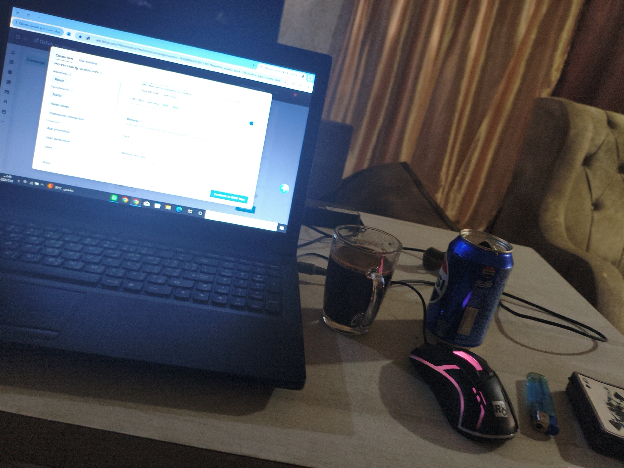 The image shows a cluttered desk with a laptop, a glass of dark liquid, a can of Pepsi, a gaming mouse, a lighter, and a card. The laptop screen displays a TikTok campaign creation interface with various options such as "Create new," "Use existing," "Awareness," "Reach," "Traffic," and "Video views." The room appears to be dimly lit, with curtains in the background. The presence of a gaming mouse and what looks like a card suggests a personal workspace. The lighting and overall atmosphere suggest it might be evening or nighttime. Based on the context provided, this scene is located in the city of Gaza, Palestinian Territories.