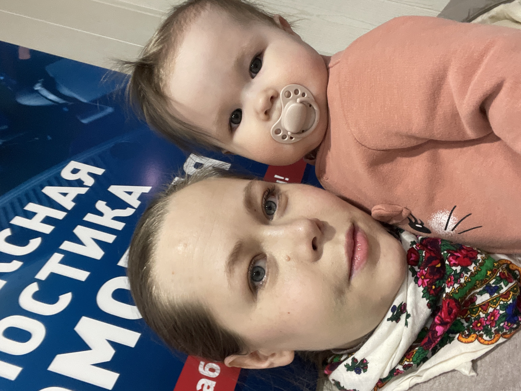 A woman and a baby lie on a blue surface with Cyrillic text on it. The baby, wearing a pink outfit, has a pacifier in its mouth. The woman is wearing a patterned headscarf. The text on the blue surface includes the words "РОССИЙСКАЯ МО" and "ав". The location is likely Chaikovsky, Russia, based on the Cyrillic text.