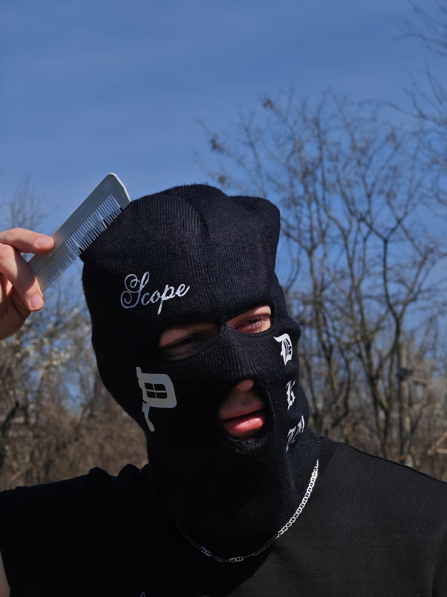 This image, captured outdoors in Shumen, Bulgaria, depicts a person from the upper torso up. The individual wears a black knitted balaclava covering the head and face, featuring white embroidered script text "Scope" across the forehead and white stylized symbols on the cheek areas. The visible eye is light brown, and the mouth is slightly open. The person's right hand, with visible light-toned skin, holds a translucent grey plastic comb, positioning its teeth against the top of the balaclava. A silver-colored chain is visible around the person's neck, resting on a black shirt. The background consists of a clear blue sky above out-of-focus, bare deciduous trees, indicating a winter or early spring season.