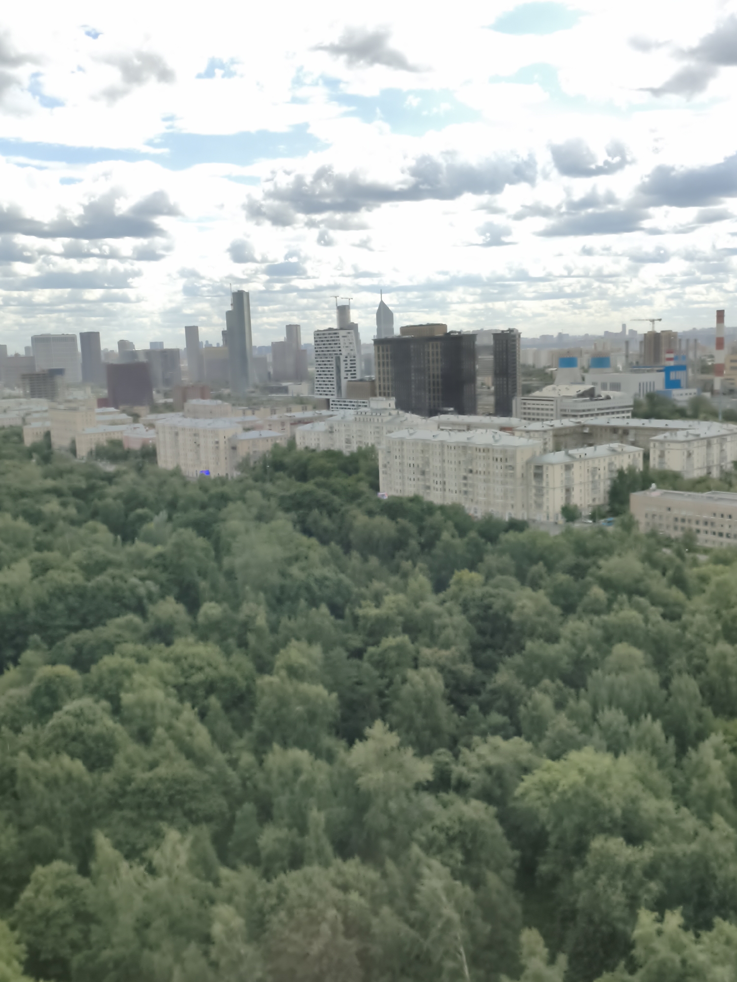 This is a high-angle, wide shot of a city skyline in Khimki, Russia, dominated by a dense forest in the foreground and a mix of residential and high-rise buildings in the background under a cloudy sky. The forest appears lush and green, with varying shades indicating different types of trees and foliage. The residential buildings are low to mid-rise, with visible windows and balconies, suggesting a suburban or established urban neighborhood. Behind these, taller, modern skyscrapers rise, some with distinctive architectural features, indicating a more central business district or developing area. The sky is partially obscured by white and gray clouds, suggesting an overcast or partially cloudy day. There are no visible people or immediate actions depicted in the scene. The overall impression is an urban environment coexisting with a significant natural green space.