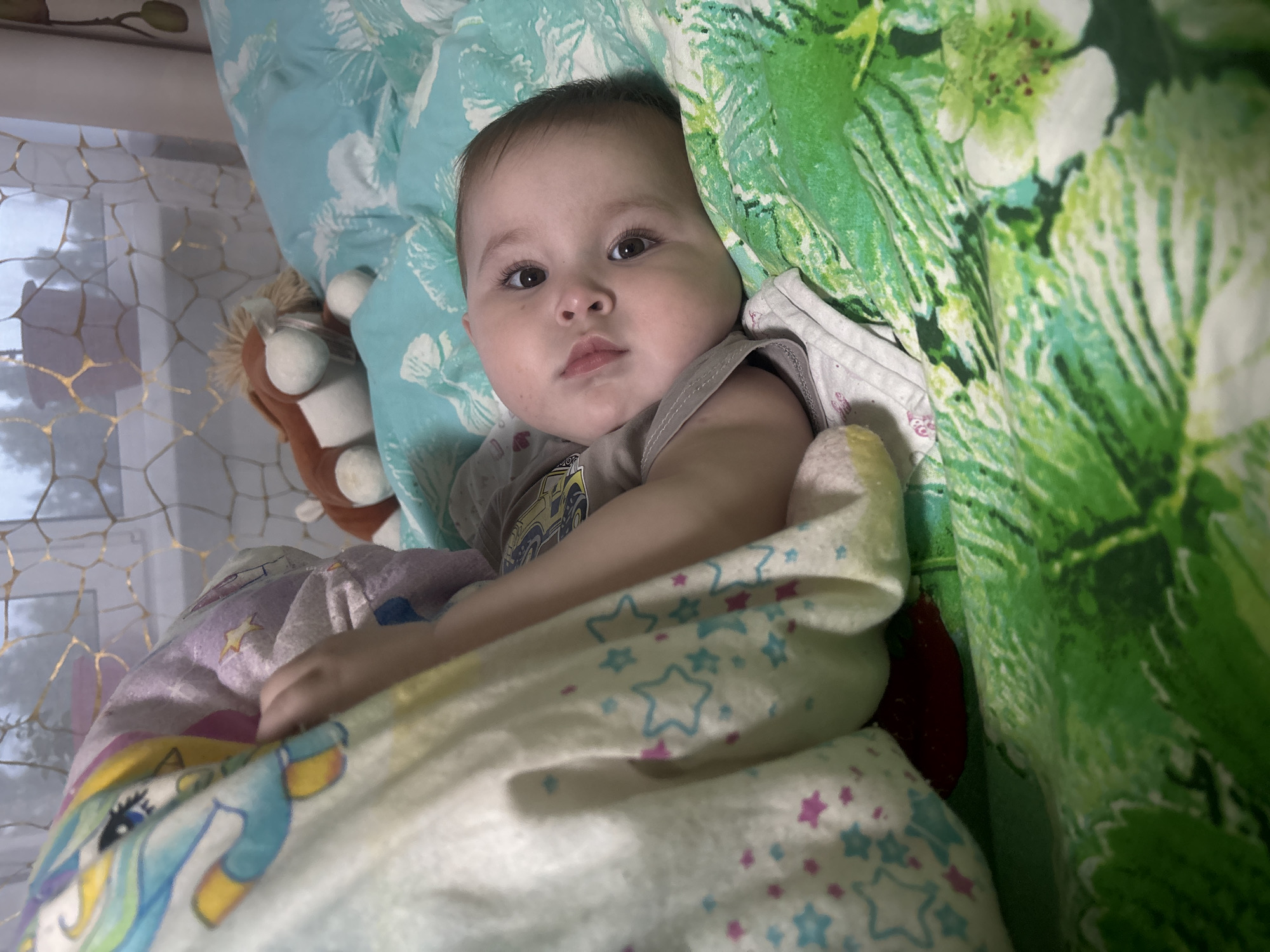An infant is depicted lying on its back on a bed, facing the viewer with a neutral expression, in the city of Chaikovsky, Russia. The baby has dark hair and brown eyes, and is wearing a light grey sleeveless top with a partially visible graphic design. Its right arm rests on a cream-colored blanket patterned with colorful stars and a stylized character. The baby's head rests on a light teal-blue pillow or duvet featuring a white floral or leaf motif. To the right and below the baby, a distinct green-patterned bedding with prominent white flowers and green leaves is visible. A light brown lion plush toy with a shaggy mane is situated on the teal bedding in the upper central-right background. Behind the bedding, a window is visible, covered by a sheer, light-colored curtain adorned with a golden, honeycomb-like pattern. A reddish-brown plant pot is discernible on the windowsill outside the curtain. A small area of red fabric is visible beneath the star-patterned blanket near the bottom center. The scene is illuminated by ambient light, suggesting an indoor domestic setting.