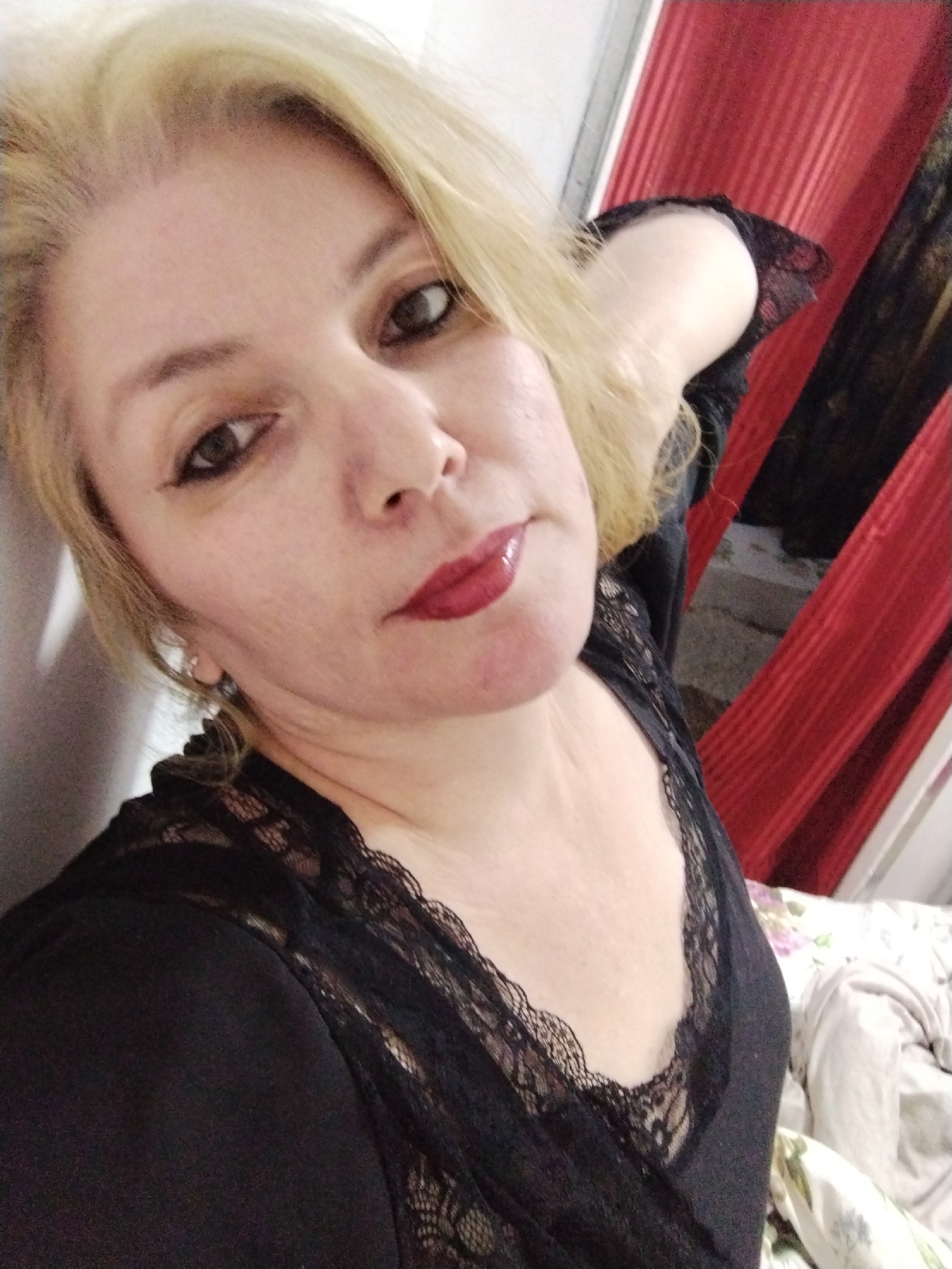 A blonde woman with light skin is posing for a selfie. She is wearing a black top with lace trim. Her left arm is raised behind her head. The background consists of a red curtain and a blurred bed with floral bedding. The image appears to be taken indoors. No specific location in Al Mas'oudi, United Arab Emirates can be determined from the image.
