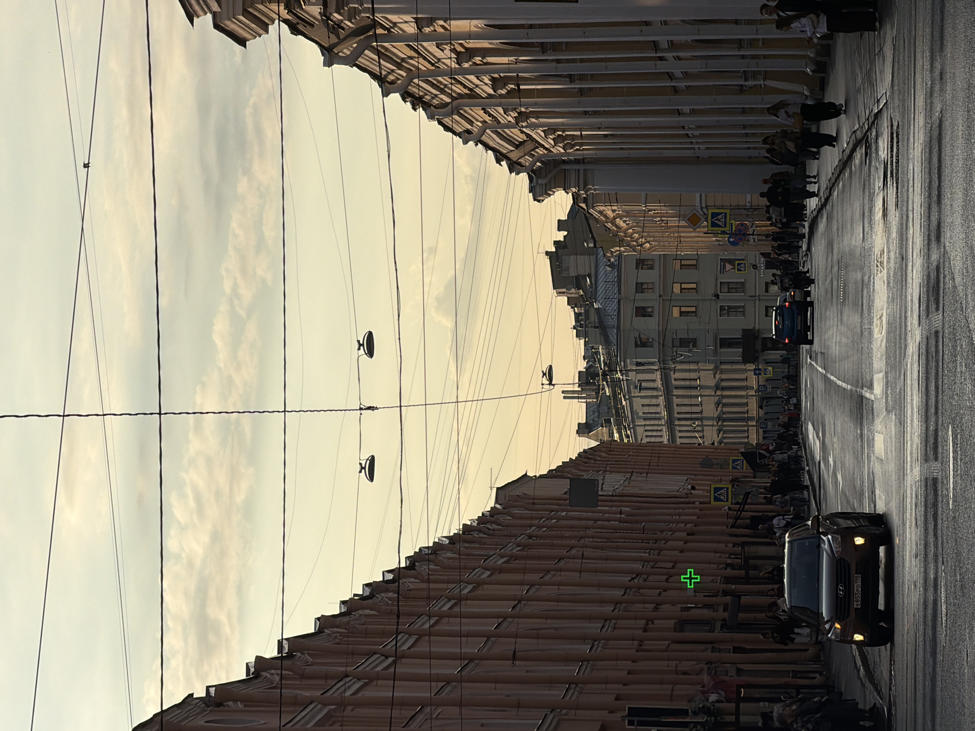 The image captures a street scene in Saint Petersburg, Russia, likely on a late afternoon or early evening given the warm, low-angled light. The perspective is from the middle of a wide asphalt road, looking down a long street flanked by tall, ornate buildings with classical architectural styles. These buildings, painted in shades of pale orange and cream, feature repetitive fenestration and decorative elements like pilasters and cornices.
Several cars are present on the road, including a dark-colored SUV in the foreground and other vehicles further down the street. Pedestrians are visible on the sidewalks on both sides of the street, some walking, others gathered in small groups. One building on the left has a bright green cross sign, indicating a pharmacy. Overhead, a complex network of electrical wires and poles crisscrosses the sky, along with a few street lamps. The sky above is a pale yellow and orange, with scattered clouds, suggesting a sunset or sunrise. The overall impression is of an urban environment with a sense of historical architecture and daily life unfolding.