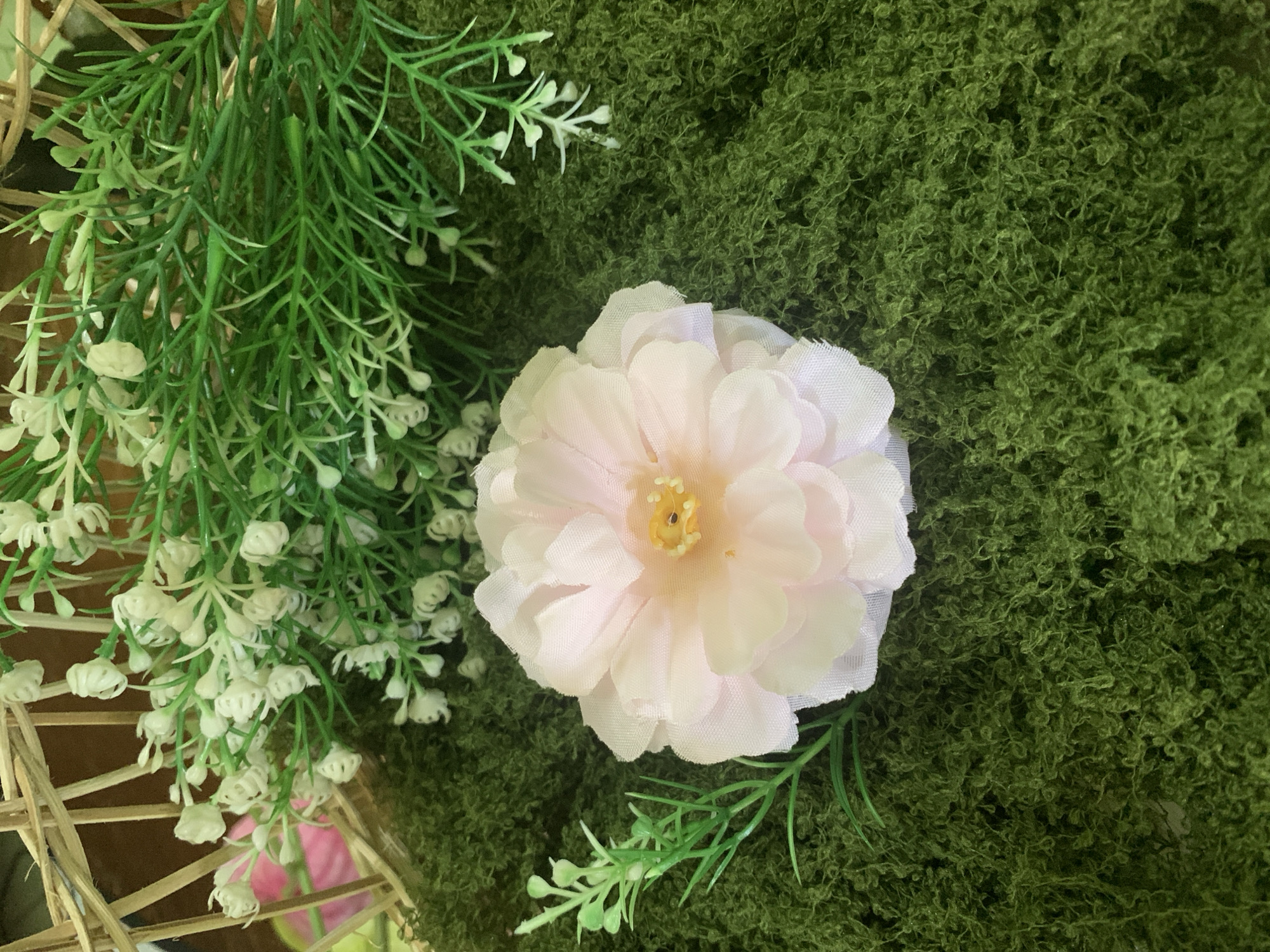 The image depicts a close-up, overhead view of a decorative arrangement. A large, pale pink artificial flower with a yellow center is positioned in the lower-middle section of the frame. It rests on a textured surface of dark green artificial moss. To the upper left and right of the flower, there are branches of artificial greenery with small white buds or flowers, and some finer, grass-like artificial foliage. A portion of a woven basket, possibly made of bamboo or wicker, is visible in the top left quadrant of the image, suggesting the arrangement is contained within or near it. The overall impression is a still-life composition of artificial floral and plant elements, likely for decorative purposes. There is no indication of any specific location in Kuala Pembuang, Indonesia, nor any people, actions, or interactions.