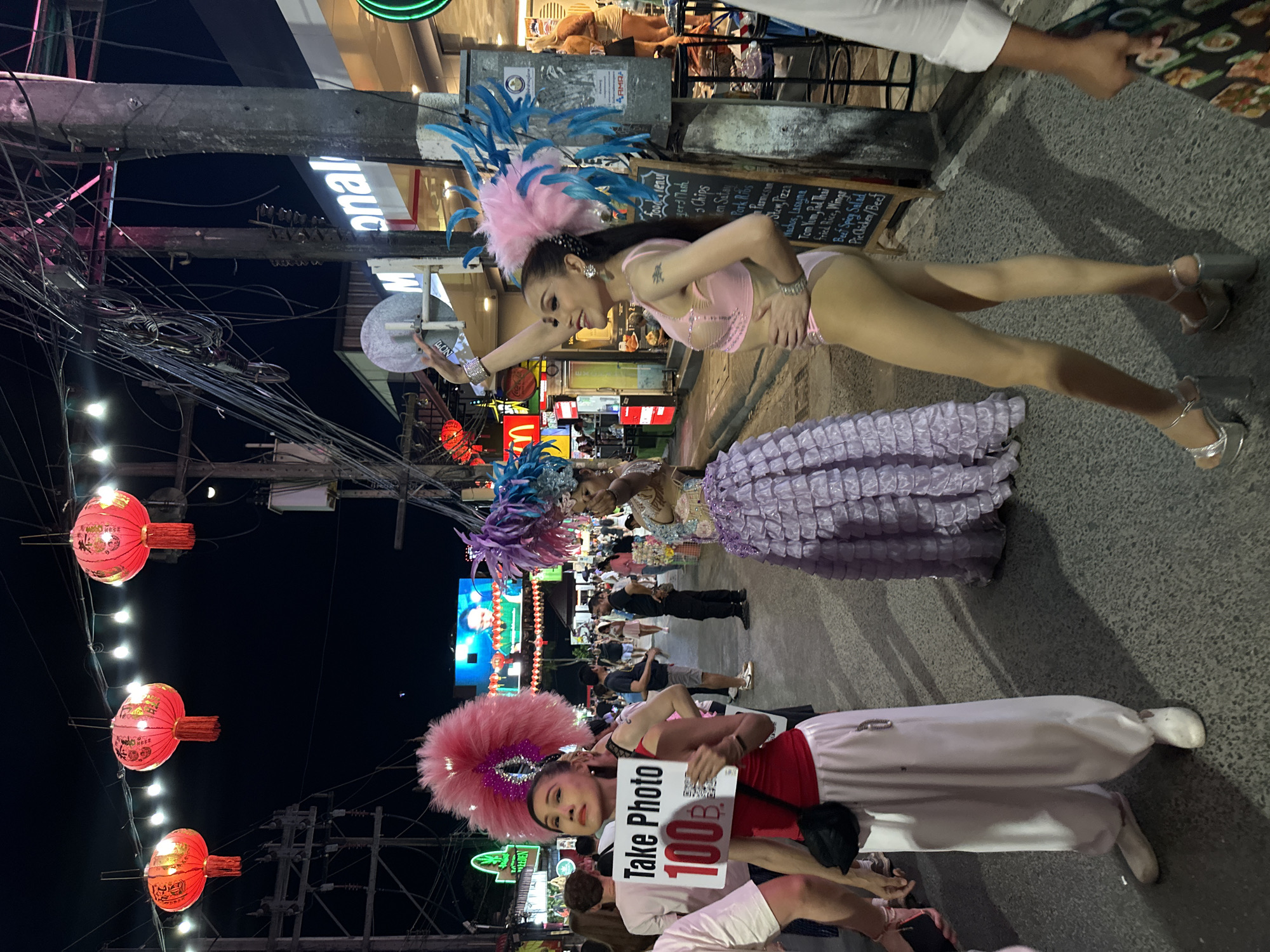 The image depicts a bustling nighttime street scene in Patong, Thailand, characterized by vibrant artificial lighting and a dense crowd of pedestrians. Three individuals, likely performers, are prominently featured in the foreground and midground, dressed in elaborate costumes.
On the left, a person with dark hair wears a red tank top, loose white pants, white slip-on shoes, and a large pink feathered headpiece. They hold a white sign reading "Take Photo 100 B." (Thai Baht) in black and red text. A small black fanny pack is slung across their chest.
In the middle, another individual wears a shimmering, ornate top and a voluminous, tiered, ruffled purple skirt, topped with a large purple feathered headpiece. Their arm is slightly raised.
On the right, a third individual with long dark hair wears a light pink sequined bra top, matching briefs/bodysuit, sheer hosiery, and silver platform high heels. They sport a pink and blue feathered headpiece. Their right arm is raised, holding a reflective silver circular object, and they are smiling towards the right.
Overhead, a string of small white lights illuminates the street, interspersed with three prominent red Chinese lanterns adorned with gold characters and red tassels. Numerous tangled black electrical and communication wires are visible against the dark sky, draped around multiple dark utility poles.
To the right, a partially obscured "McDonald's" sign is visible on a building. Below it, a black chalkboard sign displays a menu with various food items. Metal stools are present near the storefront. On the left, other commercial establishments with illuminated signs, including a green neon sign for "Cannabis," line the street.
The street surface is dark gray asphalt. Many other pedestrians in casual attire are visible in the background, walking and standing, contributing to the crowded atmosphere. The overall scene suggests a commercial and entertainment district at night.
