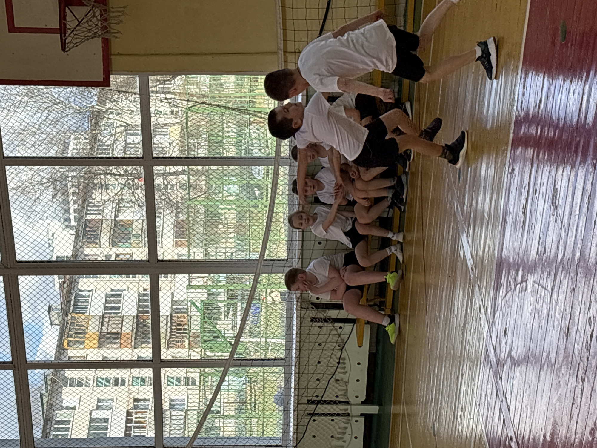 This image depicts a scene inside a gymnasium or sports hall, likely in Izhevsk, Russia. A group of young males, dressed in white t-shirts and black shorts, are present on a wooden sports court.
In the midground, several boys are seated on a long, light-colored wooden bench. One boy on the left end of the bench has his head in his hands, while others further along the bench appear to be resting or observing. To the right of the seated group, two boys are actively standing. One boy is seen in mid-stride, moving towards the right side of the frame, while another stands facing towards the left. Their postures suggest they are either participating in an exercise, playing a game, or transitioning between activities.
The setting features a wooden sports floor with visible wear and tear, including lighter and darker, possibly painted, sections. A tall sports net, likely for volleyball, runs horizontally across the middle of the hall. In the upper left corner, part of a basketball hoop with a red backboard and white net is visible.
Large windows on the left side of the hall, covered with a security grate, provide natural light. Through these windows, residential apartment buildings with multiple stories and bare trees can be seen, indicating an urban environment. The outdoor scene suggests it is daytime, and the lack of foliage on the trees points to a cooler season like late autumn or winter. There is no visible text in the image. The overall atmosphere suggests a physical education class, a sports practice, or a break during a training session.