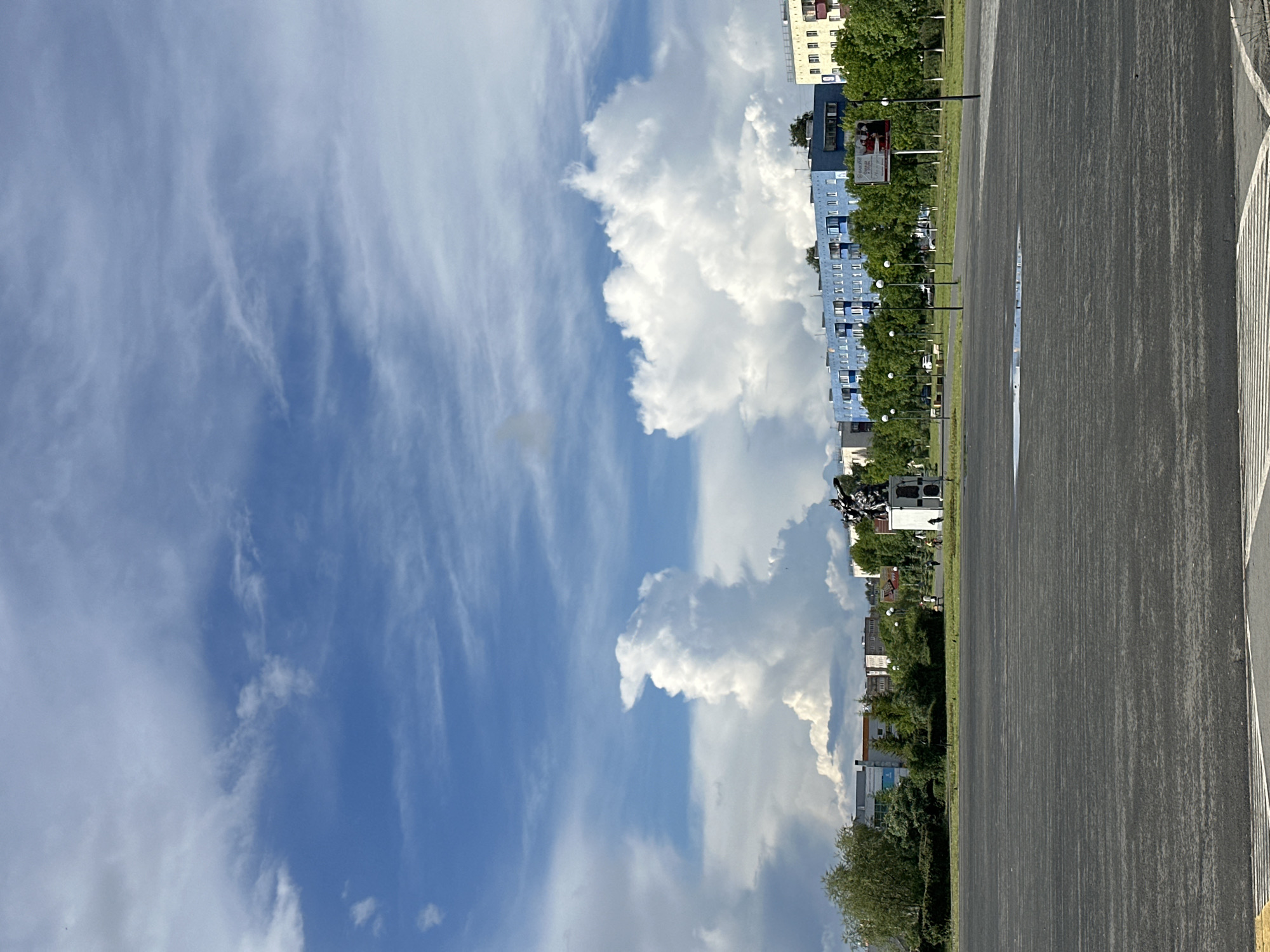 The image displays an urban or suburban landscape under a bright, partly cloudy sky. In the foreground, a wide, dark grey asphalt surface extends from the bottom of the frame towards the horizon, featuring a white painted line on its right edge. A small, reflective puddle is visible on the asphalt in the mid-ground, indicating recent moisture.
Beyond the asphalt, a strip of green grass and foliage separates the paved area from a row of buildings. Several black poles, likely streetlights or utility poles, stand along this grassy divide, receding into the distance. One of these poles carries a rectangular billboard or sign, displaying an advertisement with images of people and some barely legible text, including what appears to be "DOGANCI."
In the background, a series of multi-story buildings line the horizon. A prominent light blue building with numerous windows stands out, flanked by other structures, including one with a lighter facade. Lush green trees and bushes are interspersed among these buildings, partially obscuring them.
The sky occupies the upper portion of the frame, showcasing a vibrant blue. On the left, wispy, streaky white clouds are scattered across the blue expanse. On the right, large, fluffy white cumulus clouds are visible, suggesting fair weather. The scene is bathed in bright daylight, with no human activity directly observable.